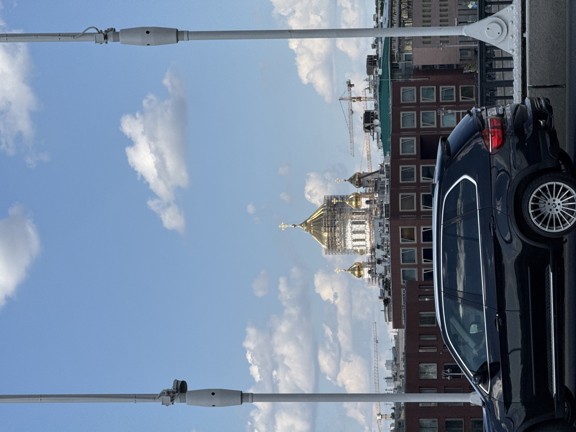 An urban scene is captured under a bright blue sky dotted with scattered white clouds. In the foreground, the rear and passenger side of a dark-colored SUV are visible, positioned on what appears to be a bridge or elevated road. Grey metal structural elements, likely part of the bridge's framework, run along the bottom and top edges of the image.
Beyond the vehicle, the cityscape unfolds with several multi-story buildings featuring reddish-brown facades and numerous windows. Some buildings have green roof sections. Dominating the mid-ground, slightly to the left of the center, are the gleaming golden domes of a large cathedral, strongly resembling the Cathedral of Christ the Savior. Scaffolding is evident around the main dome, indicating maintenance or construction.
Several tall construction cranes are scattered throughout the background, rising above the buildings and near the cathedral, signifying ongoing development. The time of day appears to be midday or afternoon due to the clear, sunny conditions. No people are visible, and there is no discernible text in the image. The overall setting depicts a vibrant urban environment in Moscow, Russia, blending historical architecture with modern infrastructure and construction.