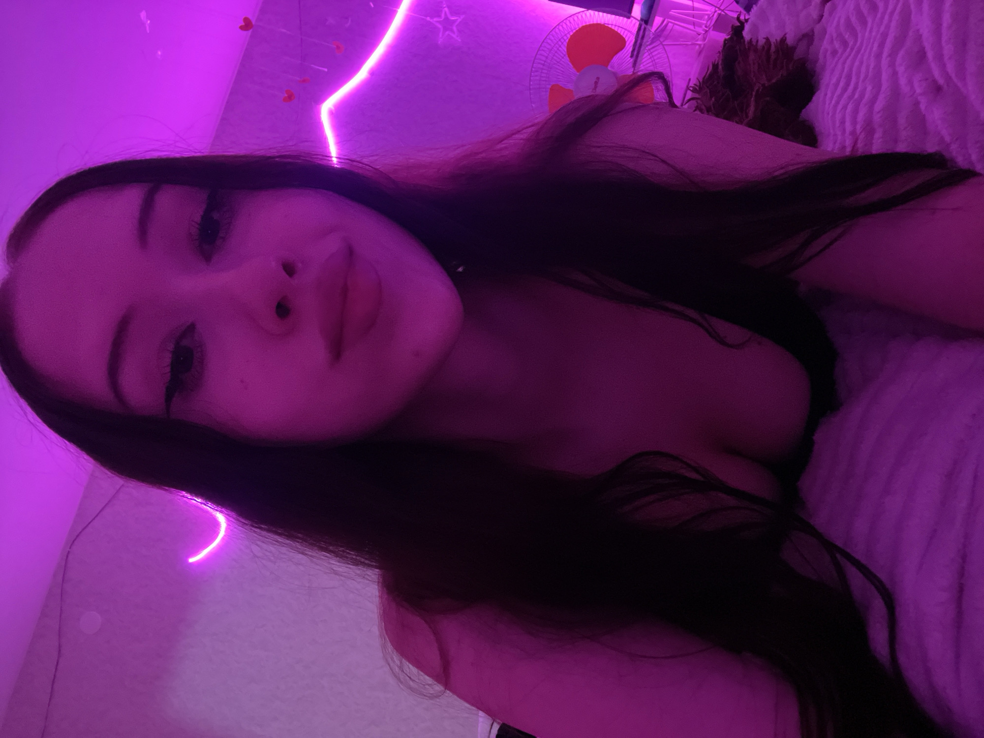 A young woman with long, dark hair lies on a bed covered with a fluffy pink blanket. She is looking at the camera, and her face is illuminated by pink neon lights. The background is also decorated with pink neon lights and small red heart-shaped cutouts. A small electric fan is visible to the right of the frame. The setting appears to be a bedroom, likely in Votkinsk, Russia.