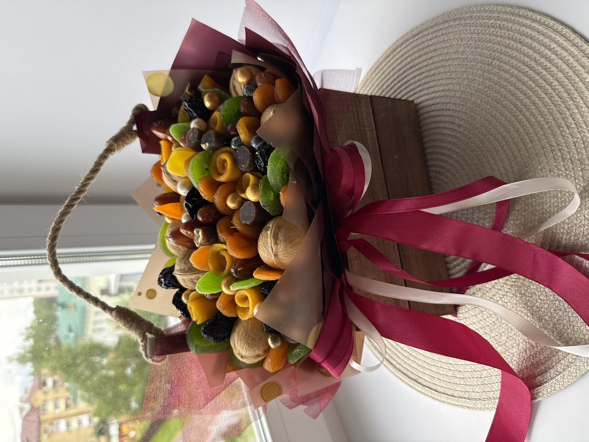 A bouquet of dried fruit is on display on a window sill. The bouquet consists of dried apricots, candied fruits, nuts, and other treats, arranged in a box with red and white ribbons tied around it. A rope handle is attached to the box. A woven straw mat is behind the bouquet. The window overlooks buildings and greenery in the city of Unknown, Unknown.