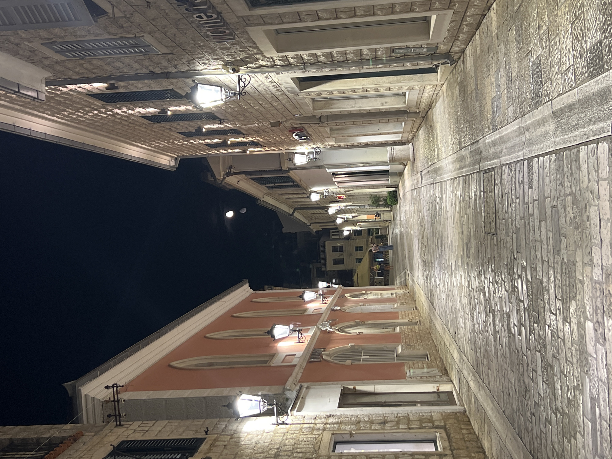 This is a night-time, eye-level, vertical shot of a cobblestone street in Herceg Novi, Montenegro. The street is lined on both sides by traditional stone buildings with warmly lit windows and decorative lanterns. The buildings on the left are painted a salmon pink with arched windows, while the buildings on the right are constructed from light-colored stone. The sky above is a deep, dark blue, with a faint moon visible. A few figures can be seen walking in the distance, suggesting the street is open to pedestrian traffic. The cobblestones of the street are wet, reflecting the light from the lanterns, indicating recent rain.