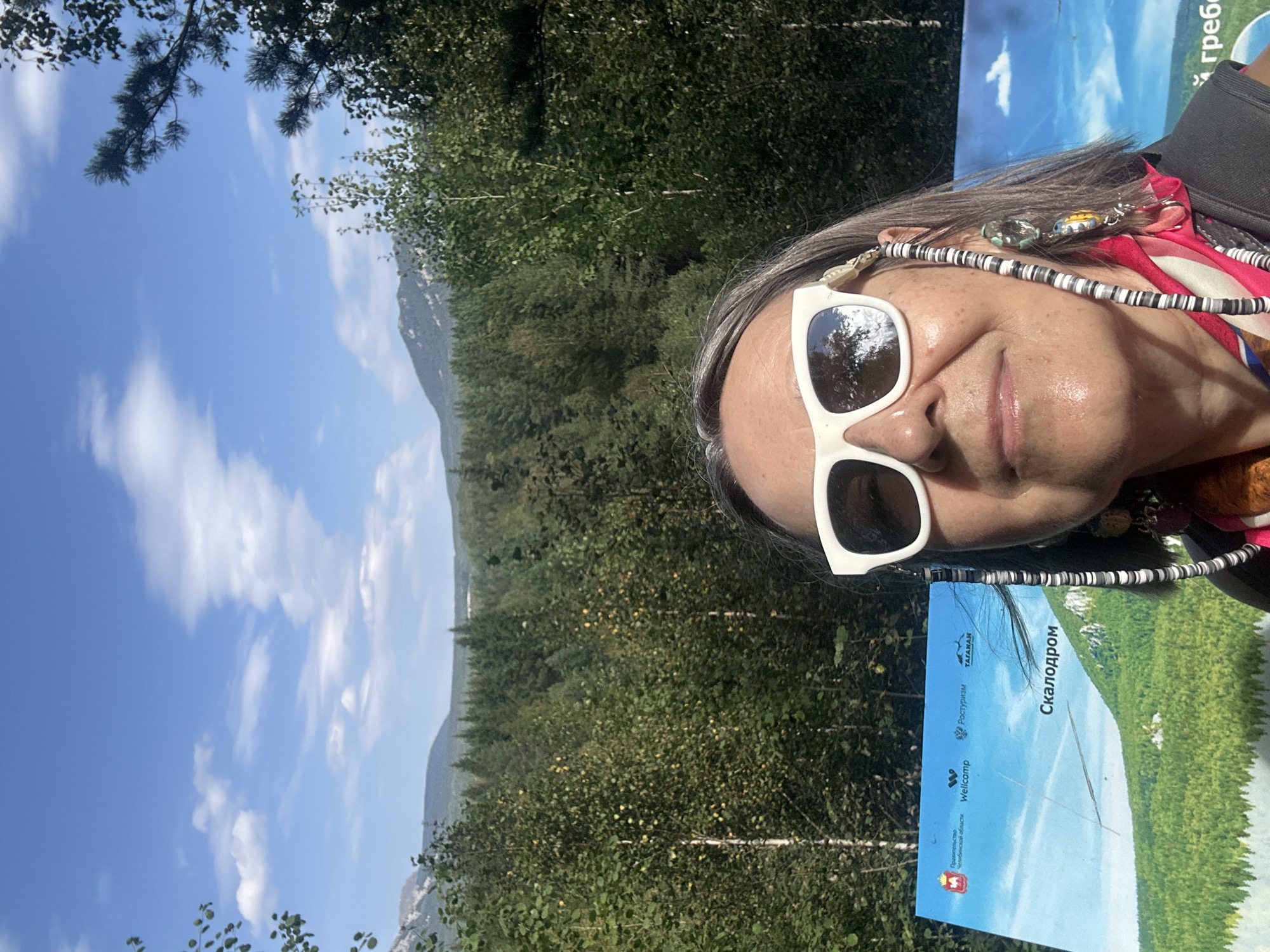 In this selfie-style photograph, a woman with grey hair and white-rimmed sunglasses is positioned in the foreground, looking towards the camera with a slight smile. She is wearing a bright pink and white patterned scarf around her neck. A beaded glasses chain with colorful charms hangs from her right lens.
Behind her, a sign is visible, featuring text in Russian and an image of a green, hilly landscape under a blue sky with clouds. The text "Скалодром" (Skaladrom) is prominently displayed on the sign, suggesting a climbing area. The background opens up to a vast expanse of green forest, leading to rolling hills and mountains in the distance. The sky above is a clear blue with scattered white clouds. The overall setting appears to be a natural, outdoor environment, likely a park or scenic area.