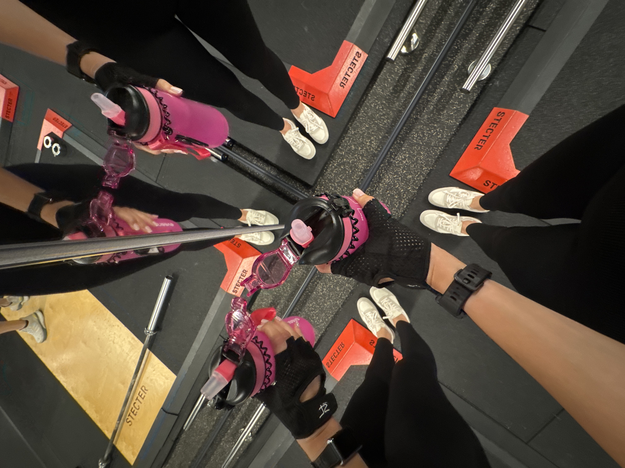 The image shows a group of people in a gym, likely a fitness class, as indicated by the matching workout attire and equipment. The subjects are all wearing black leggings and white sneakers, with several individuals also sporting black workout gloves. The focus is on their hands holding pink water bottles. The scene is shot from a high-angle, looking down at the participants and their reflections in a mirrored surface.
The activity appears to be a break or transition within a workout, with some people holding their water bottles and others resting. The gym setting is suggested by the dark, textured flooring, gym equipment like a barbell visible in the reflection, and the presence of red corner markers with the text "STECTER" printed on them. The lighting is typical of an indoor gym. No specific time of day or weather conditions can be inferred. The overall mood seems casual and focused on fitness.