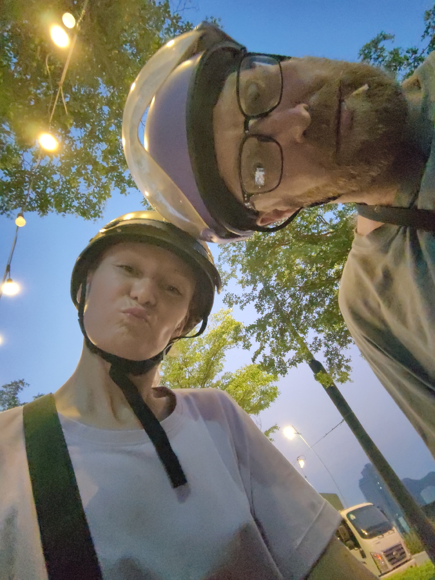 A low-angle, tilted selfie features two individuals wearing helmets. The person on the left has light skin, a light-colored t-shirt, and is making a pouting facial expression. The helmet is dark. The person on the right, presumably slightly behind, has facial hair, wears glasses, and a helmet with a transparent visor.
The background shows trees, some illuminated hanging lights, streetlights, and part of a white van. A building is barely visible in the distance. The photo appears to have been taken at dusk or dawn. The location is identified as Bai Chay Ward, Vietnam.