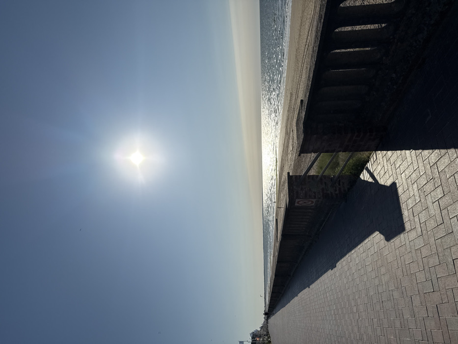A sunny seaside scene displays a hazy sky, a radiant sun, and a calm ocean. A concrete pier with shadowed stairs separates the sea from a paved walkway, creating a strong linear division. The walkway extends into the distance, lined with buildings and hinting at human presence. The lighting is bright, casting long shadows across the walkway. The horizon is blurred by atmospheric haze.