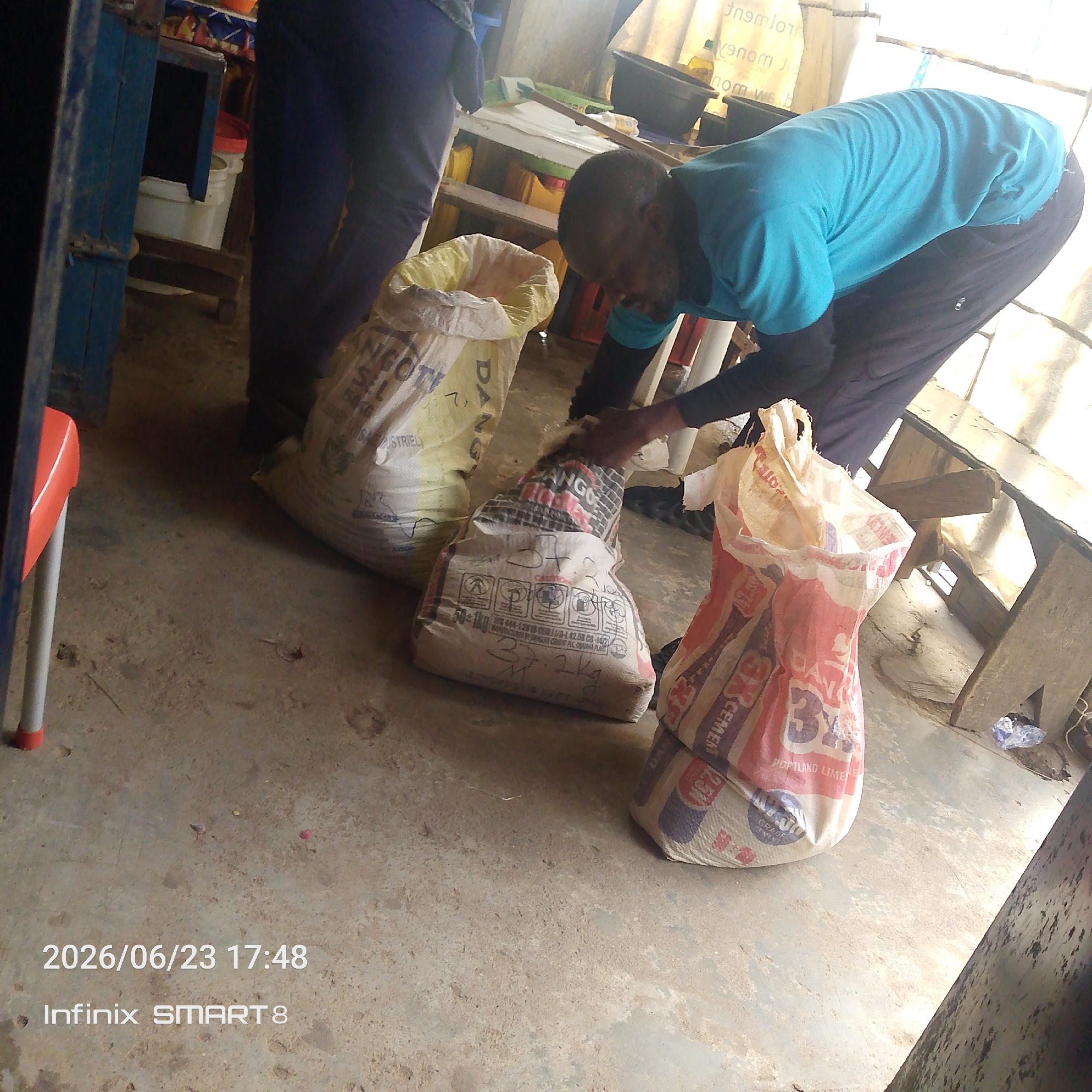 The image shows two people in what appears to be a storage area or market stall. One person, wearing a blue t-shirt and dark pants, is bent over and appears to be interacting with a bag. The other person, in dark pants, is partially visible on the left side of the frame.
Several large bags, likely containing cement or similar building materials, are on the floor. One bag has red, white, and blue branding with the text "CEMENT" and "PORTLAND LIMENE". Another bag is predominantly yellow and white with text that includes "DANGOTE STRIEL". A third bag is grey and white with a red band and what seems to be pictograms and text including "CAUTION".
The setting is rustic, with a concrete floor and wooden shelves visible in the background. There are also some buckets and other items on the shelves. The lighting suggests it could be daytime, possibly indoors or in a shaded outdoor area.
A timestamp at the bottom left of the image reads "2026/06/23 17:48" and "Infinix SMART 8", indicating the date and time the photo was taken, as well as the device used. The overall impression is of a scene involving the handling or distribution of goods.