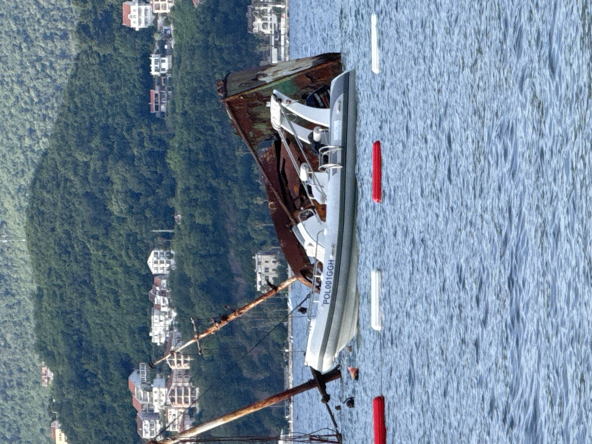 The image captures a coastal scene dominated by a partially submerged, heavily rusted vessel or large metal structure in calm, blue-grey water. In the background, densely forested hills rise steeply, populated with numerous multi-story buildings featuring white or light-colored facades and red-tiled roofs, spread across the terrain.
In the midground, the prominent rusted structure exhibits severe corrosion, displaying deep orange, reddish-brown, and some greenish oxidation. Its upper hull or superstructure and two angled, extensively rusted masts with visible rigging extend above the waterline, suggesting a state of disrepair or being a shipwreck.
Floating adjacent to the large structure is a white rigid-hulled inflatable boat (RIB) with light grey inflatable tubes. The boat features a central console, seating, a stern roll bar, and a black Yamaha outboard motor. The identification number "POL001GGH" is clearly visible on its port side.
The water surface across the foreground and midground is gently rippled. Scattered in the water are two red cylindrical buoys, three white rectangular buoys, and one black spherical buoy, along with a smaller dark object near the base of the submerged structure. No individuals are visible in the scene, which appears to be under clear daytime conditions.