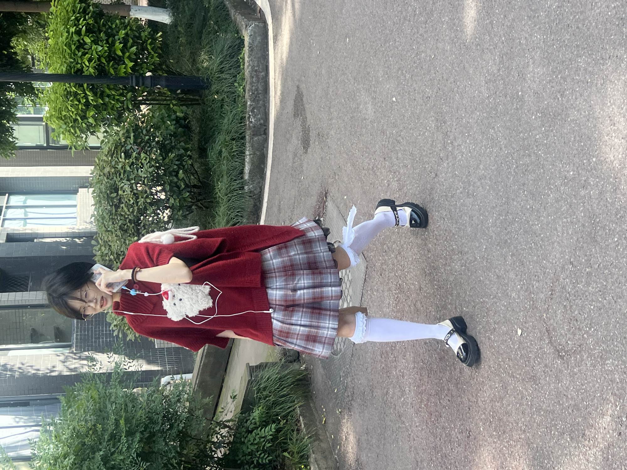 An outdoor, slightly rotated full-length shot shows a young person walking on a paved path in Jiangjin, China, on a sunny day. The individual has short, dark hair and wears glasses, a loose-fitting red sweater, a red and white plaid skirt, white knee-high socks with ruffled tops, and black and white chunky platform shoes. They are holding a white smartphone to their right ear, engaged in a conversation, with white wired earphones connected. A small, white furry plush toy (possibly a dog or rabbit) hangs around their neck on a white strap, positioned near their chest. A white backpack or bag is partially visible on their back.
To the left of the path, there is a concrete curb bordering an area of green grass and lush bushes. Behind the foliage, glimpses of modern buildings with rectangular windows can be seen, suggesting an urban or campus environment. The path itself is dark gray asphalt or concrete, showing subtle variations in texture and some lighter patches, possibly from sunlight or wear. Shadows are visible on the ground, indicating bright daylight. No visible text is present in the image.