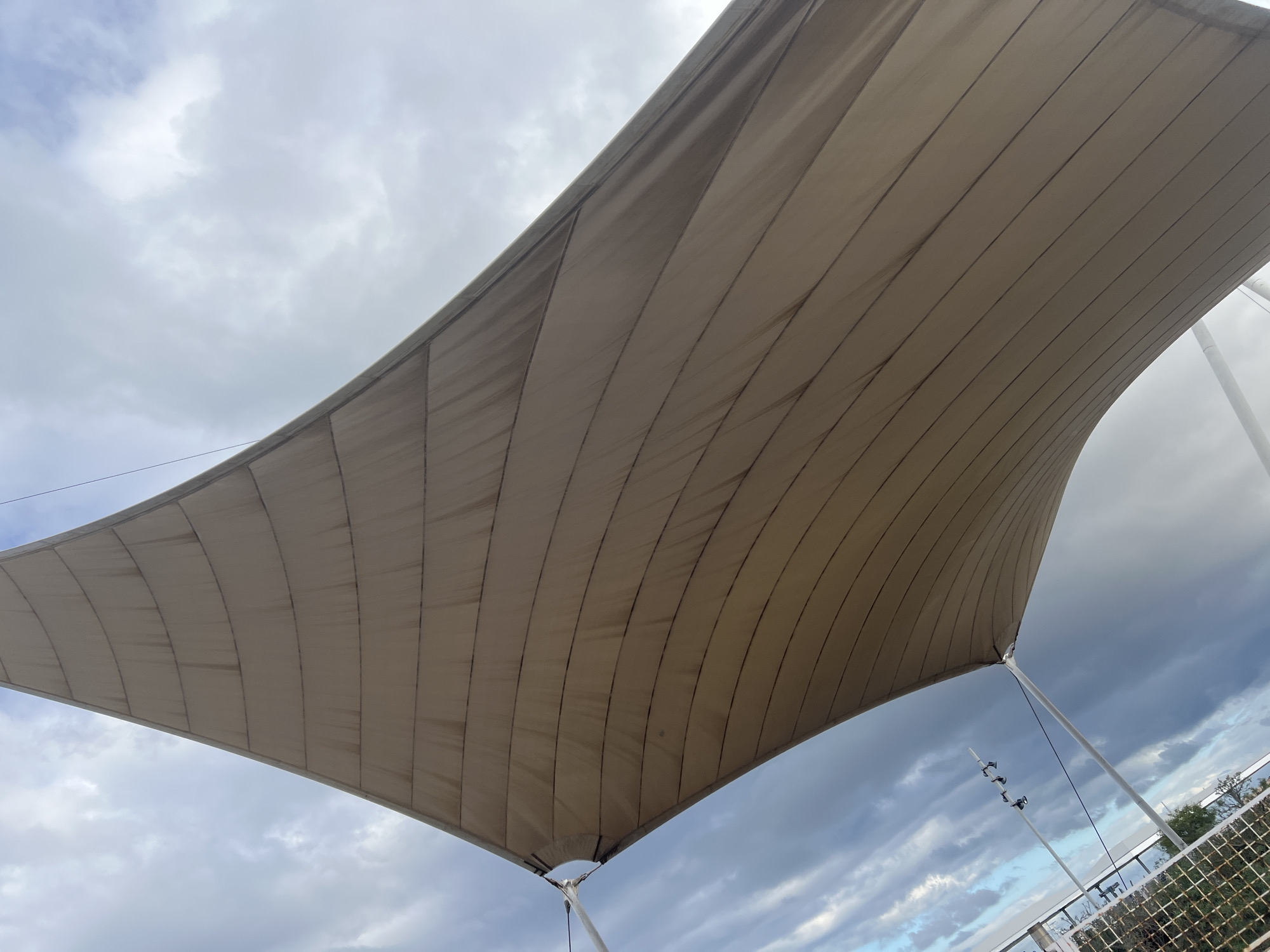 The image captures a large, light-colored tensile fabric canopy from a low-angle perspective, looking upwards. The canopy, likely a shade structure, is a muted beige or off-white and is composed of multiple rectangular panels stitched together, forming distinct parallel seams that curve gracefully across its surface. These seams show subtle signs of weathering or dirt.
The fabric structure is supported by slender, white poles at its corners, with visible tension wires or cables extending from the fabric to these supports. One such pole is prominent in the lower right, along with several small, dark fixtures attached to it, possibly lights or sensors.
The background is dominated by a cloudy sky. The clouds exhibit varying shades of grey, from bright, diffused white in some areas to darker, more dramatic formations in others, indicating an overcast day with potentially changeable weather conditions. In the lower right quadrant, beyond the canopy's support, a white metal grid fence or railing is partially visible, behind which appears some green foliage, suggesting bushes or trees, and distant, indistinct structures.
The scene depicts an outdoor architectural installation in a public or semi-public space. Based on the provided location context, this structure is situated in Las Palmas, Spain, likely providing shade in a sunny environment. The time of day is daytime, under cloudy conditions. There are no people present, no specific activity taking place, and no visible text.