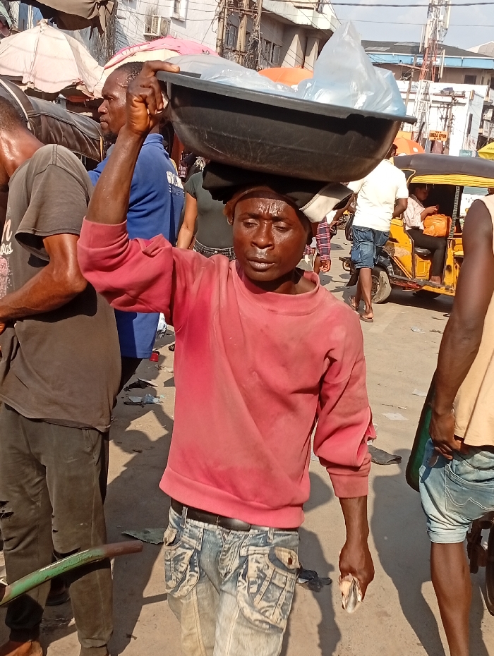 A man carries a large black tray filled with white items on his head, navigating a crowded outdoor market. He wears a sweat-stained red long-sleeved shirt and faded, cargo-pocketed jeans. The setting includes pedestrians, vendors, and a yellow vehicle in the background. The scene appears to be busy and bustling, reflecting typical market activity.