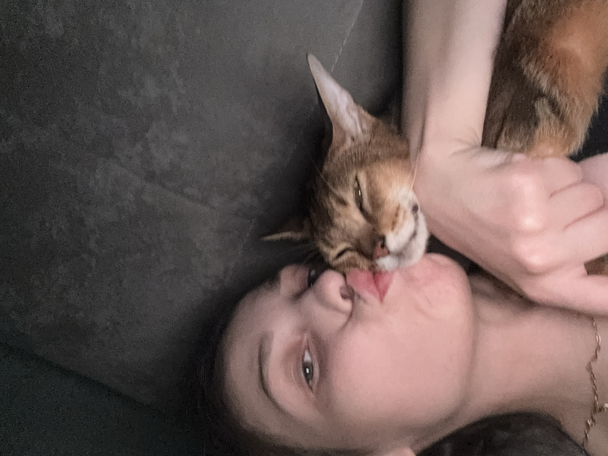 The image, taken indoors in Noviy, Russia, shows a woman and a cat. The woman is positioned on the left side of the frame, with her face turned towards the cat and her lips puckered in a kissing gesture. Her left eye is partially visible and appears to be looking at the camera. The cat, an Abyssinian breed, is on the right side, nestled against the woman. Its eyes are partially closed, and its tongue is extended, touching the woman's lips. The cat's fur is a reddish-brown tabby pattern. The background is dark and appears to be a textured surface, possibly a sofa or cushion. The lighting is dim, suggesting an indoor setting, likely a home environment.