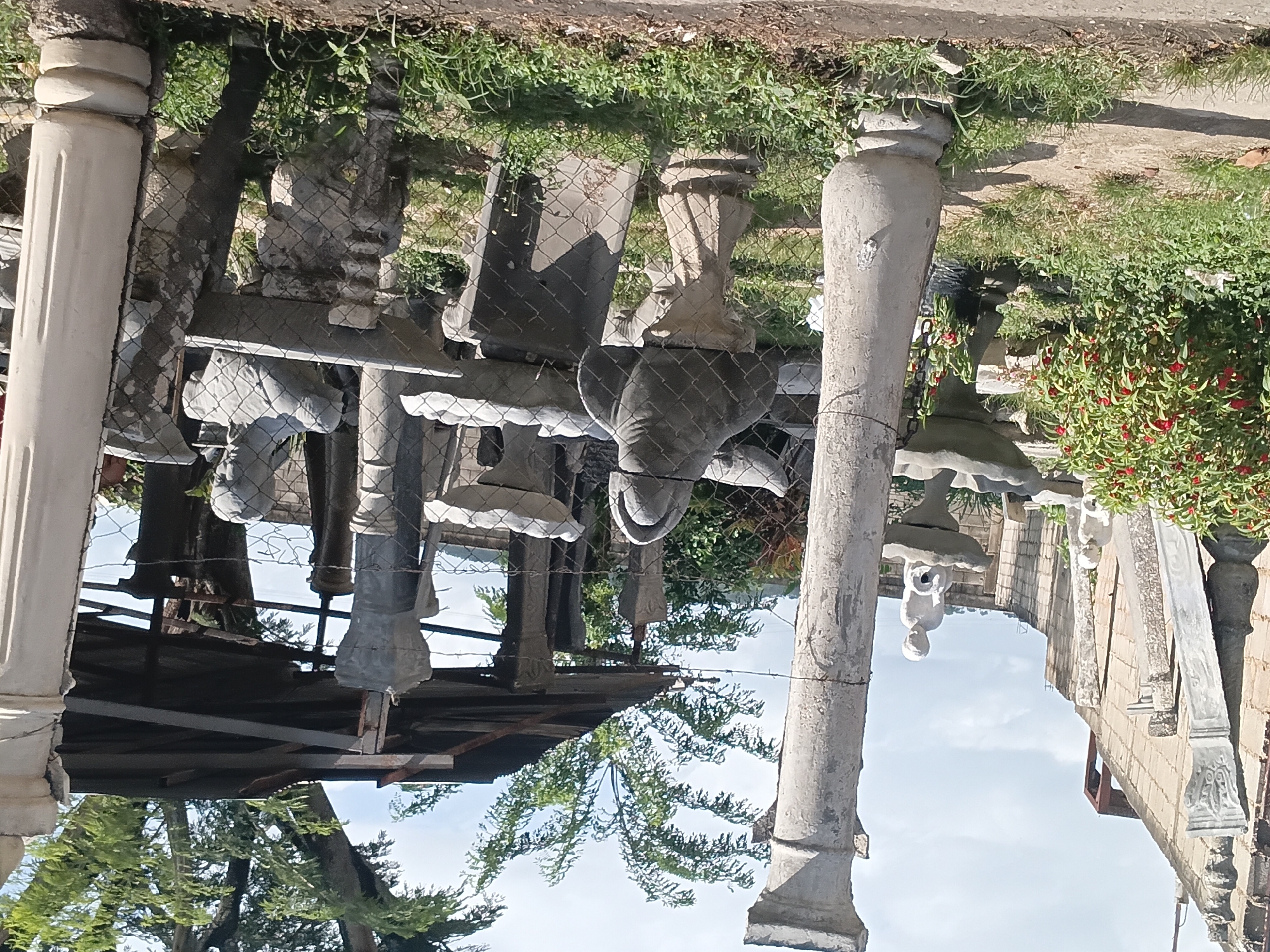 This is a ground-level, eye-level, medium shot of an outdoor display of concrete statues and architectural elements, possibly for sale, in Taguanes, Venezuela. The scene is cluttered with various items, including several classical-style columns, some of which are tall and fluted, while others are shorter and decorative.
Dominating the foreground are several statues. To the left, a large, partially obscured fluted column stands prominently. In the middle ground, a statue of a child holding a jug sits atop a fountain base, and to its right, another statue of a child is visible. Further to the right, a bust of a person and a fountain basin are present. In the background, a covered area with a corrugated metal roof supported by metal beams shelters more statues and columns.
The environment consists of overgrown grass and a dirt pathway in the foreground, suggesting an unkempt or working yard. A chain-link fence runs through the scene, separating different areas and partially obscuring some of the statues. A dense bush with red flowers is visible to the left, and large trees with green foliage can be seen behind the covered structure. The sky above is partly cloudy. The overall impression is one of a yard or storage area filled with decorative concrete items.