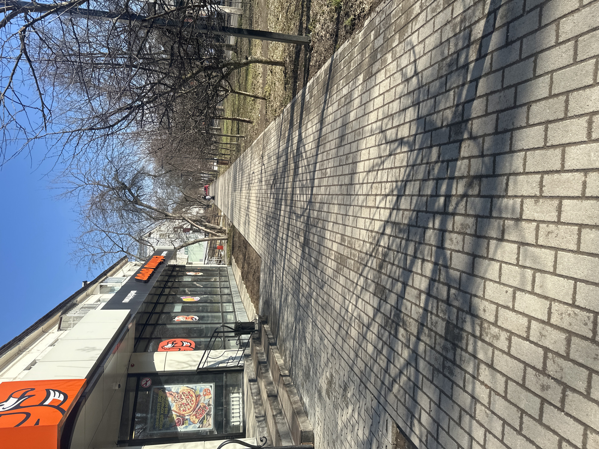 A street view in Chaikovsky, Russia, shows a long, paved sidewalk leading into the distance, marked with the shadows of bare trees. A commercial building stands to the left with signage reading "Додо Пицца" (Dodo Pizza). No people are directly visible, and the environment is indicative of early spring, with bare trees and patchy grass.