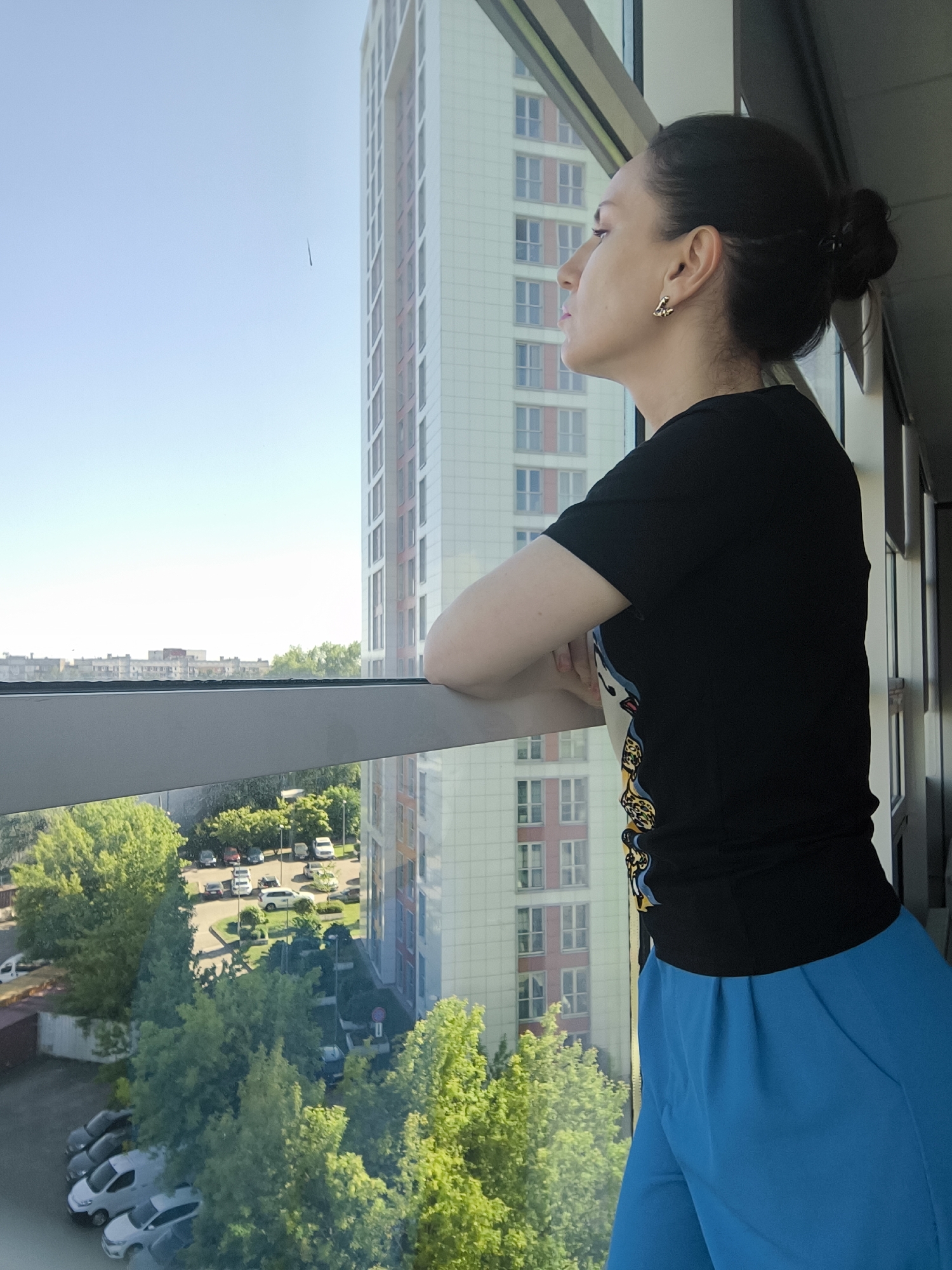 A woman is depicted in a side profile, looking thoughtfully out of a large window. Her dark hair is styled in a bun at the back of her head. She wears a black t-shirt with a colorful, possibly animal-print or patterned design featuring yellow and blue elements on the side, and bright blue pleated pants. A gold-colored earring shaped like the letter 'E' is visible in her right ear. Her forearms are crossed and resting on the window sill as she gazes upwards and outwards.
The scene outside the window reveals a clear, bright blue sky, indicating a sunny day. To the right, a modern, multi-story building with a grid pattern of windows and a light-colored facade rises high. In the middle ground, a horizontal window frame separates the sky and distant buildings from the immediate outdoor view below. This lower section features numerous vibrant green trees, a parking lot with several parked cars, and sections of road. Other buildings are visible in the distance beneath the horizon. The overall impression is an urban or suburban setting viewed from a high vantage point during the day.
No other explicit text is visible apart from the earring.
Location context: Unknown, Unknown.