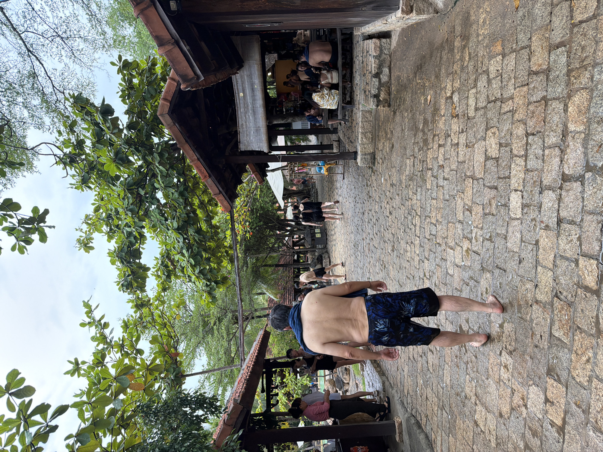 In Nha Trang, Vietnam, this image captures an outdoor scene within a resort or park area, characterized by a cobblestone pathway and traditional Southeast Asian architecture. The pathway is wet, suggesting recent rain or a watering system. Several structures with dark wood and red-tiled roofs are visible, providing shade. Lush green trees with large leaves overhang the area.
In the foreground, a shirtless man with dark hair is walking away from the camera on the wet cobblestone path. He is wearing dark blue patterned swim trunks and has a dark blue towel draped around his neck.
Other people are present in the background, some seated at wooden tables under a covered pavilion, others standing or walking. One group consists of individuals at a table, appearing to be dining or socializing. Another group is standing near a building with a red roof. To the left, a woman in a pink shirt is interacting with other individuals. The overall atmosphere suggests a relaxed, possibly recreational setting.