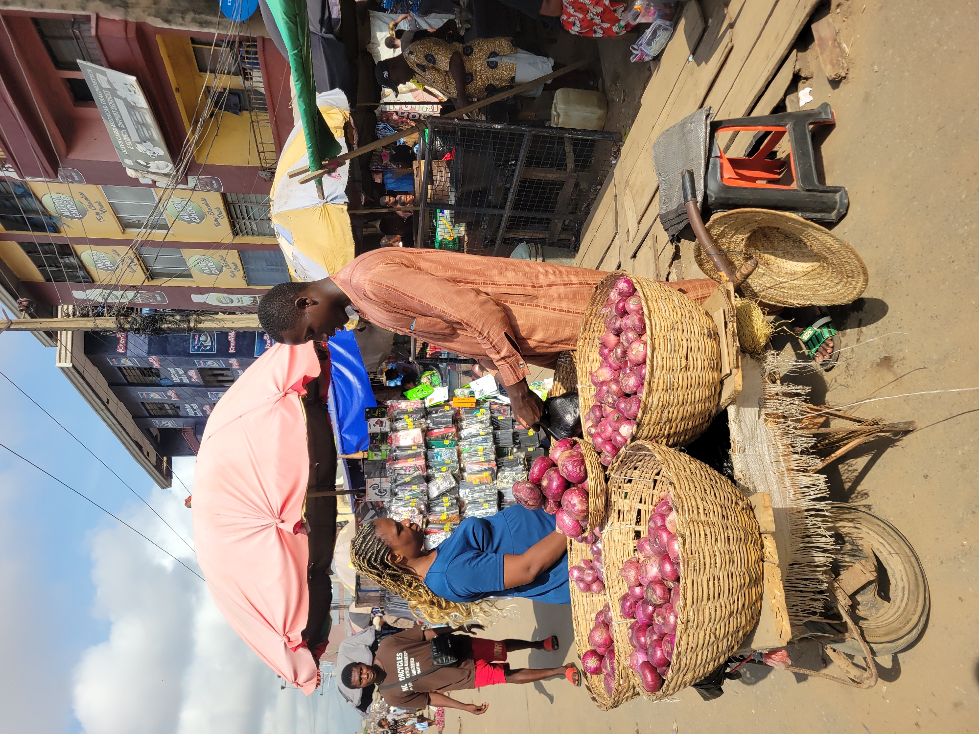 In Fegge, Nigeria, the image captures an outdoor market scene. A man in a brown striped shirt is interacting with a woman in a blue shirt, both standing near a cart laden with two large woven baskets filled with red onions. The cart also holds a straw hat and various other items. A pink umbrella provides shade for the scene. In the background, another market stall displays items for sale, including phone cases. A building with visible advertising is behind the stalls, and pedestrians are seen walking in the foreground. The ground appears to be a paved surface, typical of a market area.