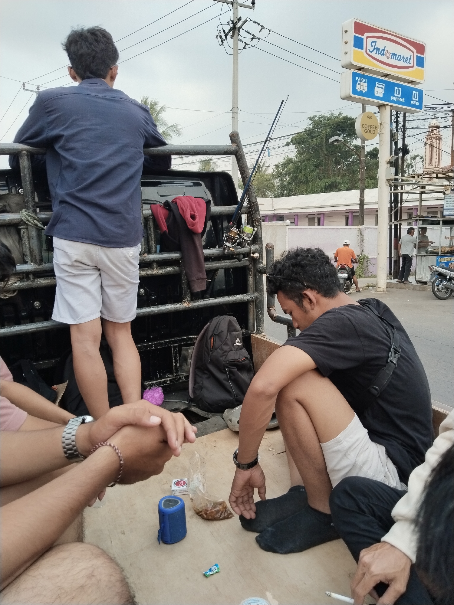In the city of Sukanagara, Indonesia, a group of young men are gathered in the back of a pick-up truck. The scene appears to be near a street, with a building and a prominent Indomaret convenience store sign in the background. The sky is overcast.
One man, seen from behind, wears a navy blue shirt and light-colored shorts. He leans on the truck's railing, looking outwards. To his right, a backpack rests against the truck's side.
Another man is seated in the truck bed, facing forward and looking down. He wears a black t-shirt and light-colored shorts, with black socks. His left arm is bent, and his hand rests on his knee. A black strap from his t-shirt is visible.
In the foreground, another man's hands are clasped. He wears a watch on his left wrist and a woven bracelet on his right. Nearby, on the truck bed, are a small blue speaker, a packet of cigarettes, and a clear plastic bag containing food items. A purple object, possibly a ball, is also visible.
A third man is partially visible in the lower right of the frame, wearing dark pants and a light-colored hoodie. His hands are visible, one holding a lit cigarette.
In the background, a man on a motorcycle wearing an orange shirt rides away from the camera. Another man stands near a food stall in front of the Indomaret. Fishing rods are propped up against the truck. The overall atmosphere suggests a casual gathering, possibly after an activity or on the way to one.