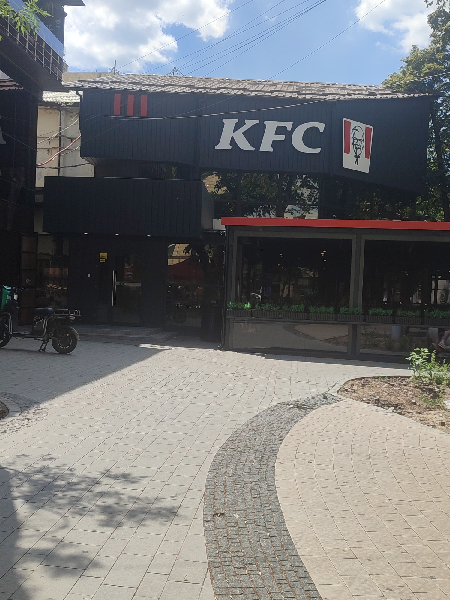 The image shows the exterior of a KFC restaurant located in Chernivtsi, Ukraine. The KFC sign is prominently displayed on the building, featuring the brand's logo. In front of the restaurant, there is a paved walkway with a curved design. To the left of the walkway, a scooter with a green delivery box is parked. The building has large glass windows, and the area is surrounded by trees and a blue sky with scattered clouds. The scene appears to be an urban environment on a sunny day.