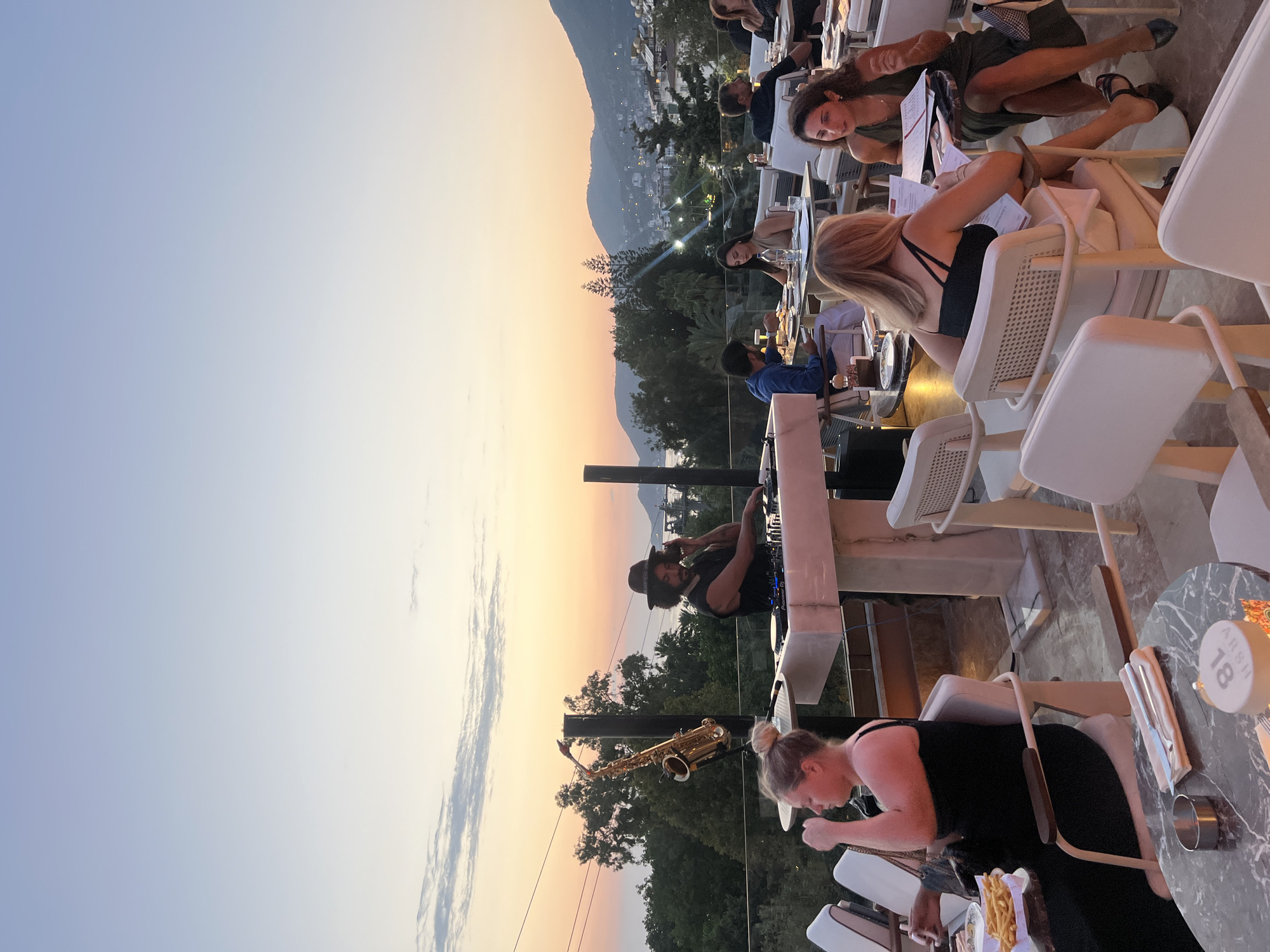 This is a wide shot of an outdoor rooftop restaurant in Alanya, Turkey, during sunset. The sky exhibits a gradient of soft blues and oranges. In the foreground, several tables are occupied by diners, mostly women, engaged in conversation or looking at menus. A DJ is set up on a raised platform, operating turntables, with a saxophone propped beside him. The background features lush green trees and the distant cityscape of Alanya, with buildings cascading down hillsides towards the sea. The overall atmosphere is relaxed and social, with a live music element.