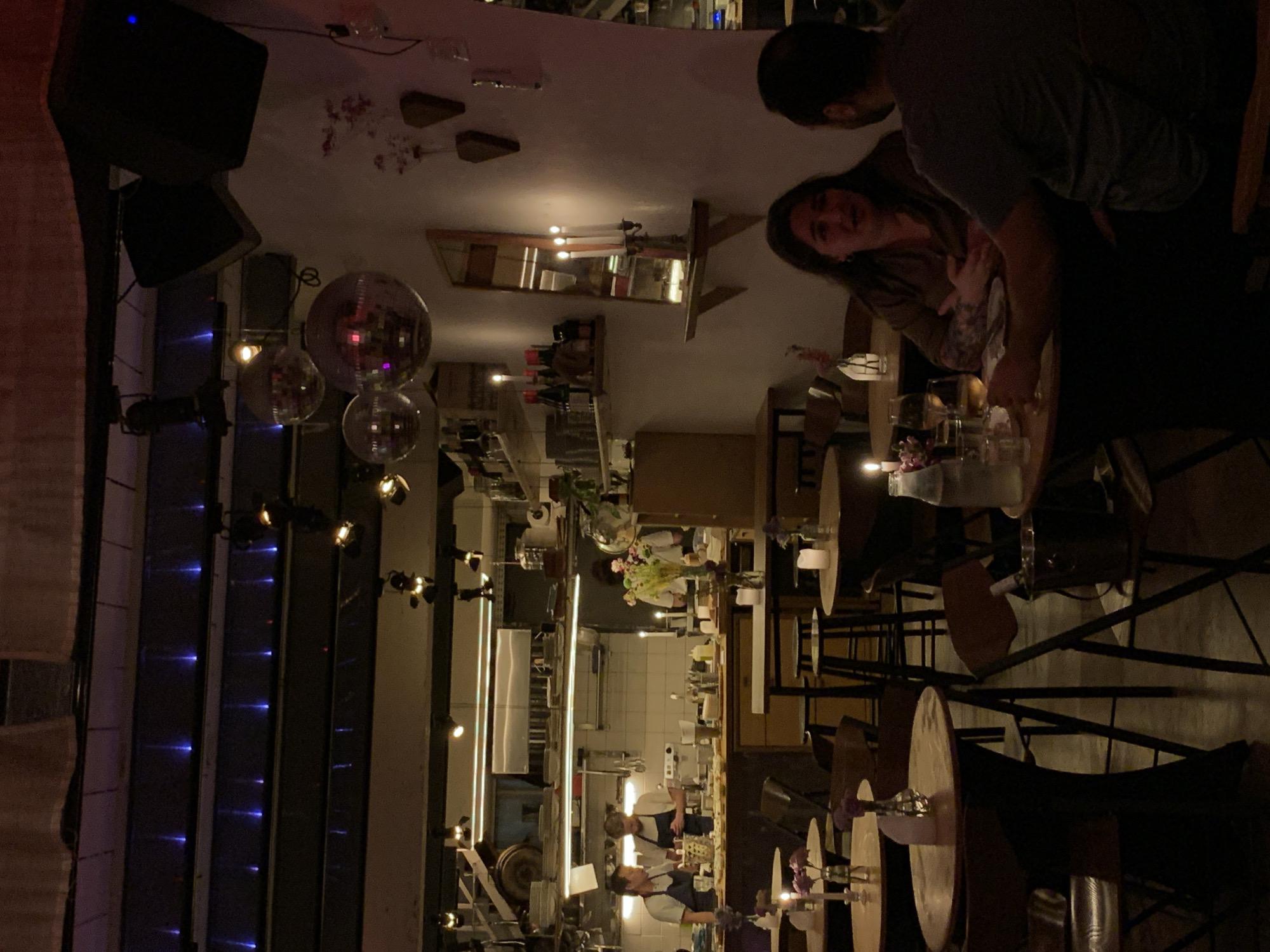 This image captures the interior of a lively establishment, likely a restaurant or bar, during the evening or night. The overall ambiance is dim and warm, with various artificial light sources creating an inviting atmosphere.
In the foreground, a man and a woman are seated at a wooden table. The woman, on the left, is seen smiling towards the camera, while the man, facing away, is partially visible. Their table, along with several others, is set with what appears to be drinks and small flower arrangements, illuminated by candlelight.
The setting includes multiple wooden-topped tables and dark metal chairs scattered across the floor. On the left side of the scene, large black speakers are mounted, and several reflective disco balls hang from the ceiling, suggesting a dynamic atmosphere or potential for music and dancing. A section of a wall features prominent vertical blue LED lights, adding a cool contrast to the warm lighting elsewhere.
In the mid-ground and background, an open kitchen or bar area is visible. Several staff members, including at least two wearing aprons, are seen working behind a counter with various kitchen equipment, including stainless steel surfaces. A long, rectangular mirror adorned with candles is mounted on a wall in this area, reflecting some of the interior.
The activity suggests people are dining and socializing, while staff are busy preparing food or drinks. The dim lighting, disco balls, and open kitchen concept contribute to a modern and casual, yet vibrant, urban dining or entertainment experience. No discernible text is visible in the image. The location is Tel-Aviv, Israel.