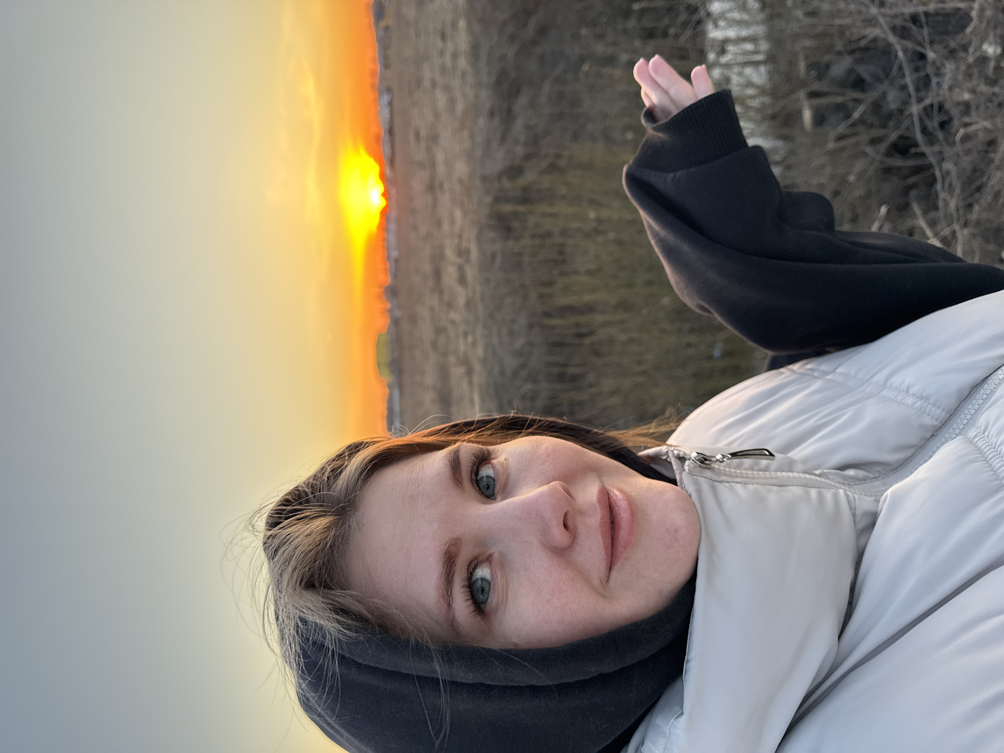 A young woman with fair skin, blue eyes, and light brown hair stands outdoors during a sunset. She wears a black hooded sweatshirt under a light gray puffer jacket. Her hand is raised slightly. The background shows a field and a setting sun. The location is Горный, Russia.