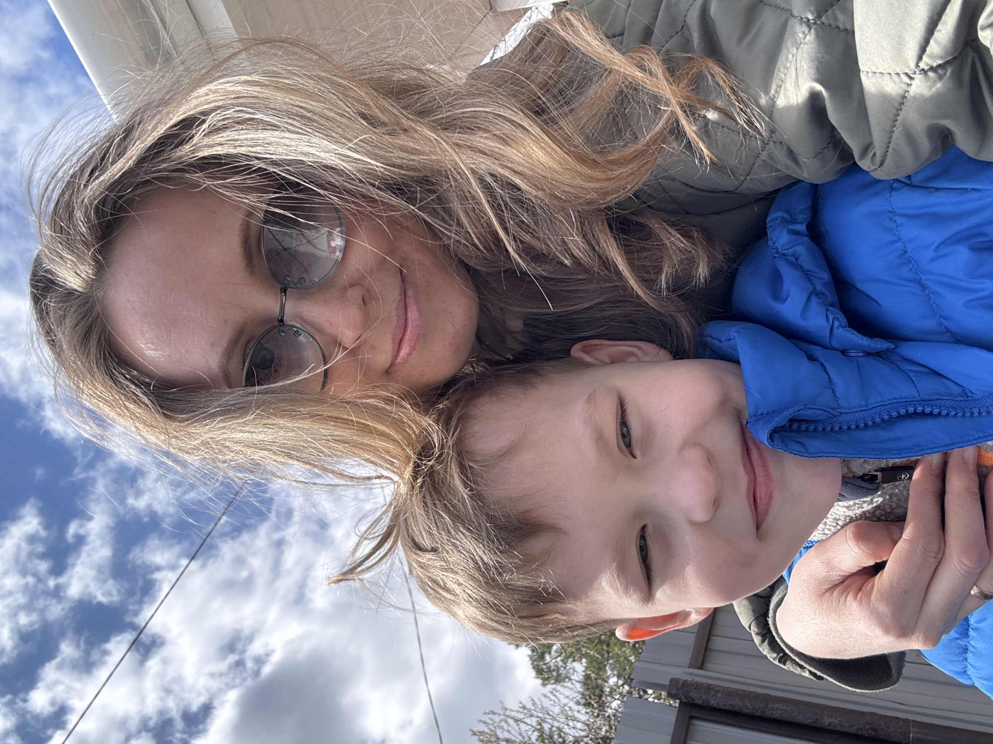 A woman and a young boy are posing for a selfie. The woman has blonde hair and is wearing glasses and a quilted jacket. The boy is wearing a blue jacket. They are outside on a cloudy day. The location is unknown and cannot be determined as Chaikovsky, Russia.