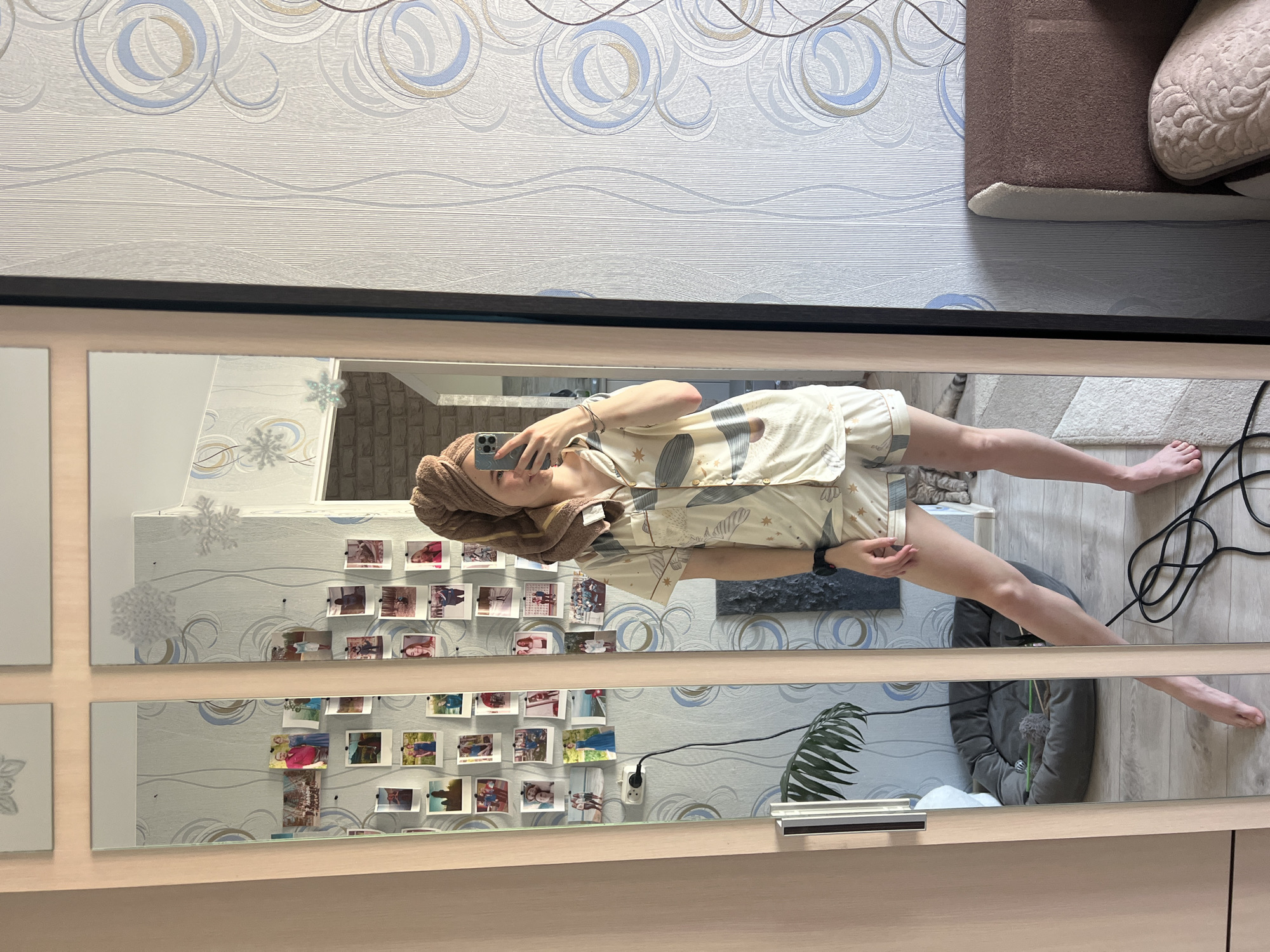 A person is taking a mirror selfie in what appears to be a room in Chaikovsky, Russia. The person has a towel wrapped around their head and is wearing pajamas. The mirror reflects a wall decorated with photos. The reflection also shows furniture and electrical cords on the floor.