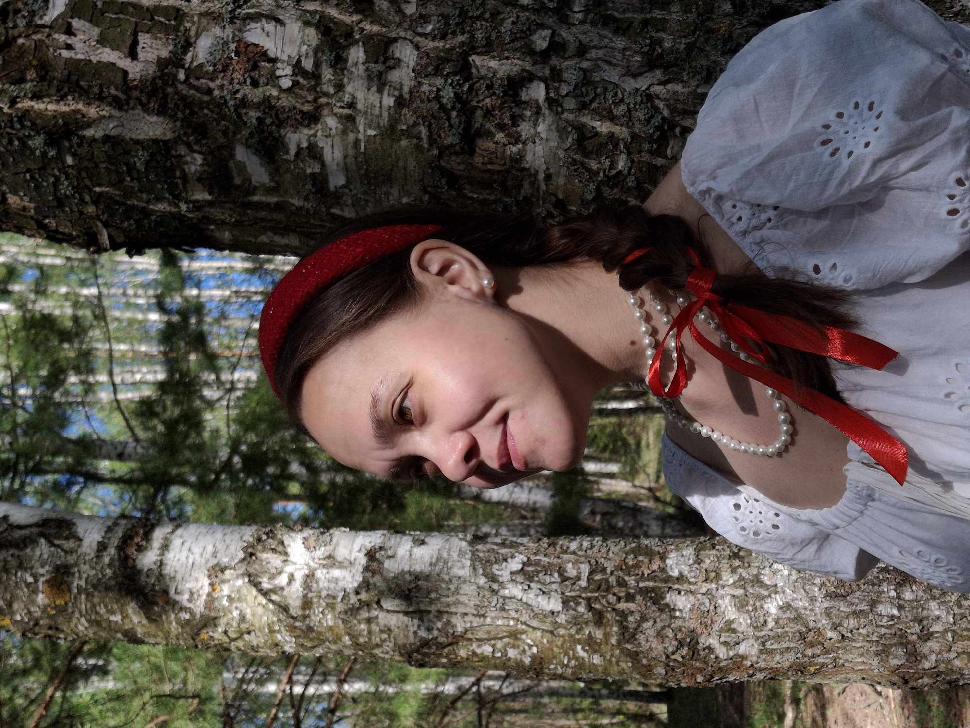 A woman with a red headband and a pearl necklace is posing near a birch tree in a wooded area. She is wearing a white blouse with eyelet detailing and a red ribbon in her hair. The tree's bark is textured with white and grey markings. The background shows more trees and foliage under dappled light, suggesting an outdoor location. The city location is Chaikovsky, Russia.
