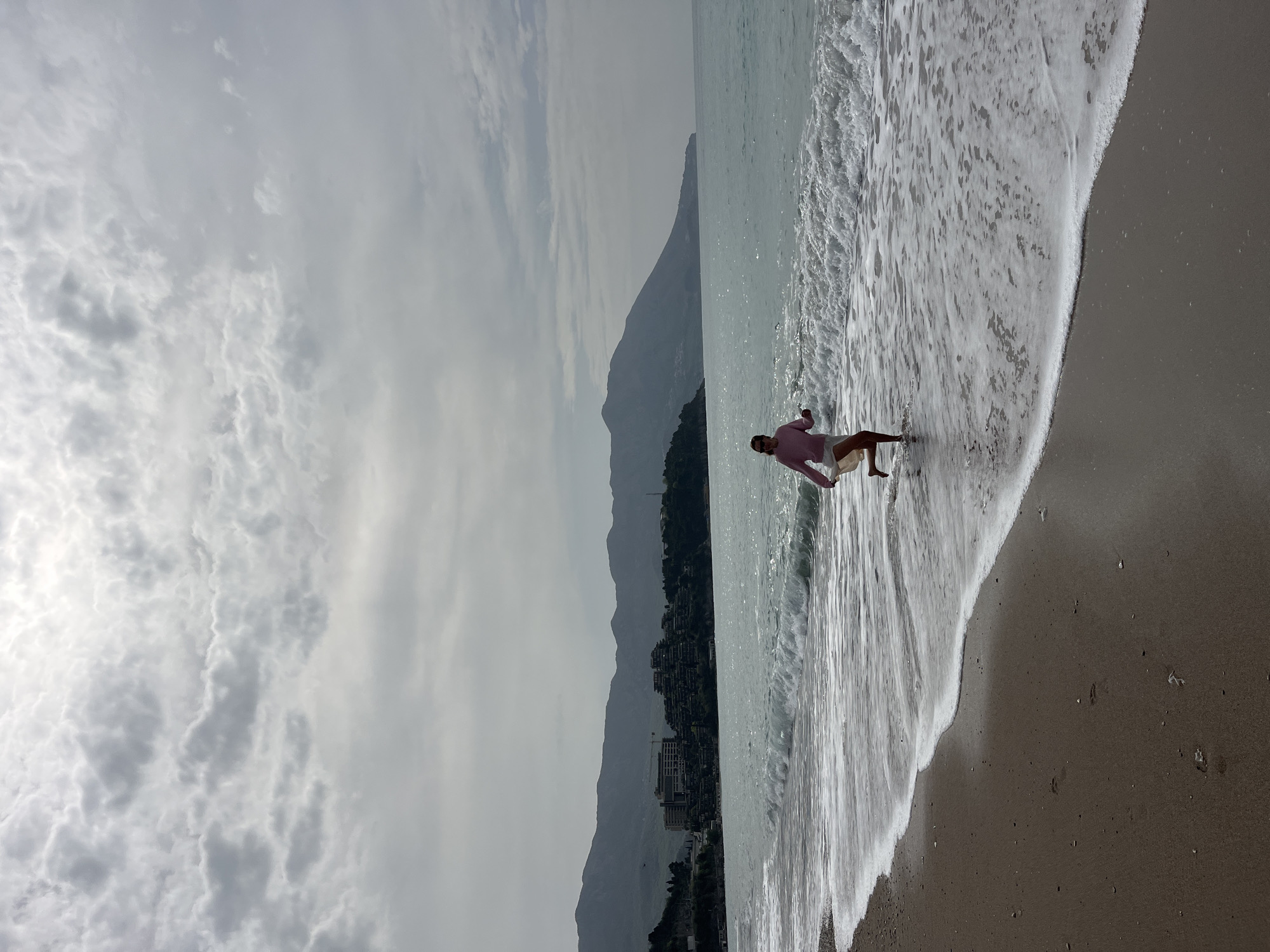 The image shows a beach scene with a person standing in the water. The beach has dark sand and is lapped by foamy waves. The person is wearing a pink shirt and white shorts and appears to be walking in the water. In the background, there are mountains and buildings, indicating a coastal city. The sky is overcast with cloudy patterns. Based on the architectural style of the buildings and the mountain range, the location is likely Budva, Montenegro.