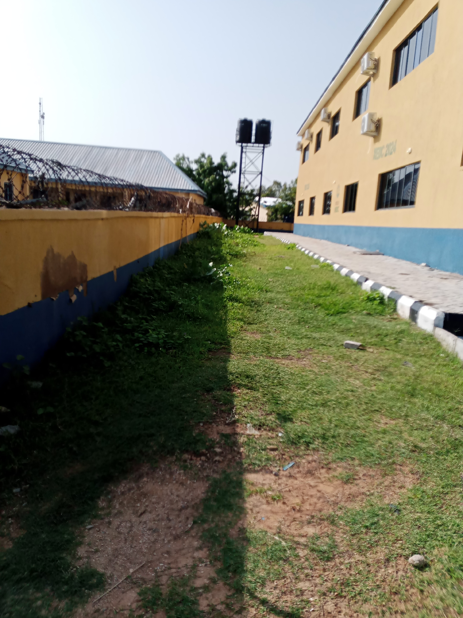 The image depicts an outdoor scene in Jalingo, Nigeria, focusing on the exterior of a building and its surrounding grounds. On the right side of the frame is a two-story building with a light yellow facade and a blue trim along the bottom. The building features multiple windows with dark frames and reflective glass. Several air conditioning units are mounted on the exterior walls. The words "NESIC 2024" are visible in green lettering on the side of the building.
To the left of the building, there is a paved walkway bordered by a decorative curb with alternating black and white sections. Adjacent to the walkway is a strip of grass that transitions into a dirt path with patches of grass and some scattered debris.
Further to the left, a yellow wall topped with barbed wire fencing encloses an area. Behind this wall, the roof of another building with a corrugated metal surface is visible, along with some trees and what appears to be a telecommunications tower with antennas. In the background, the sky is clear and bright. The scene is illuminated by daylight, casting shadows from the vegetation onto the ground.