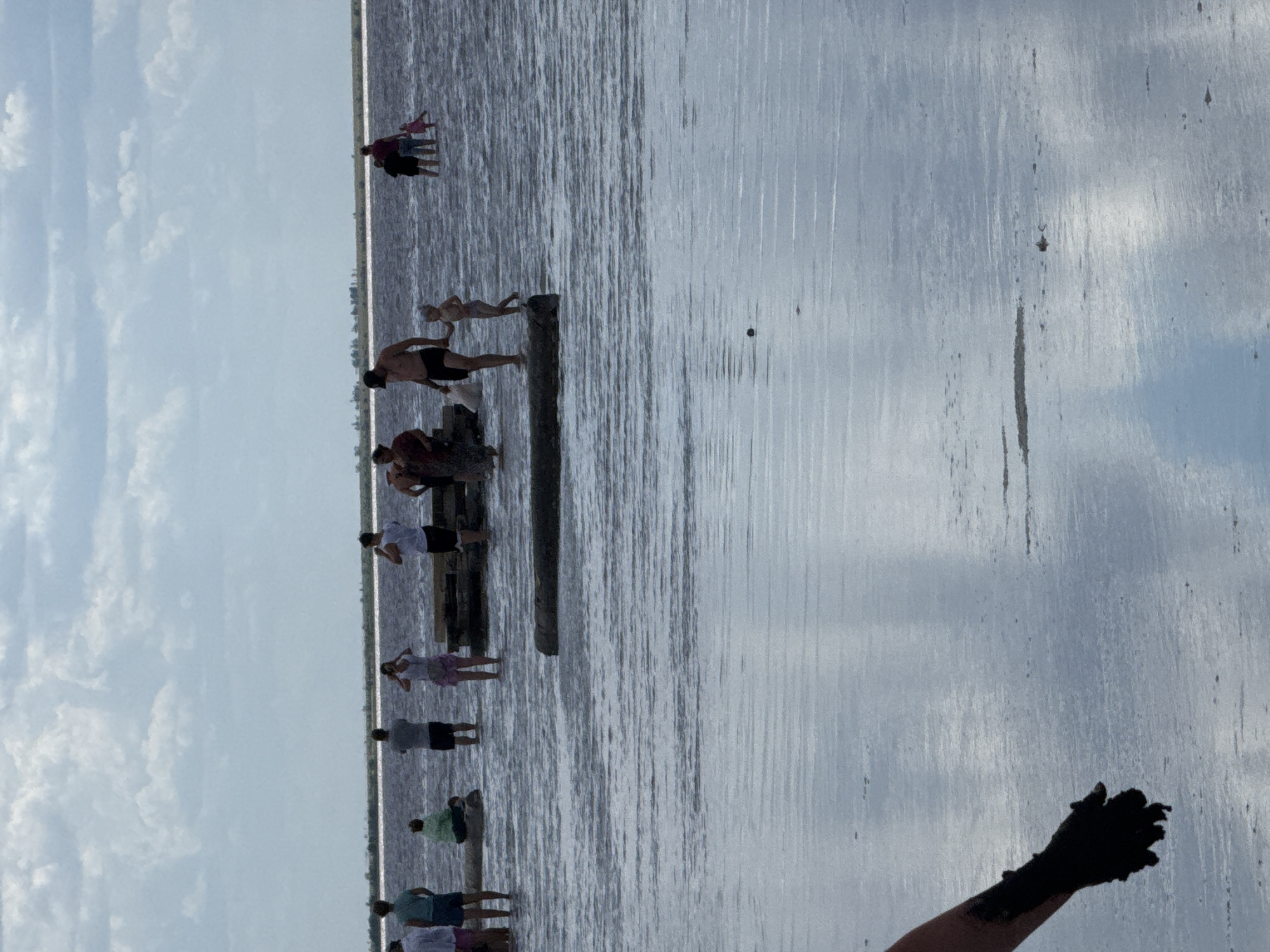 In the city of Бурсоль, Russia, this image captures a group of people wading in a shallow body of water, likely a salt lake, under a cloudy sky. The water's surface reflects the sky and surrounding landscape. Several individuals are visible in the mid-ground, some standing in the water, others on or near submerged logs. A person's arm, covered in dark mud, is prominently featured in the foreground, suggesting the practice of mud bathing. The overall scene depicts a recreational activity in a natural, possibly therapeutic, environment.