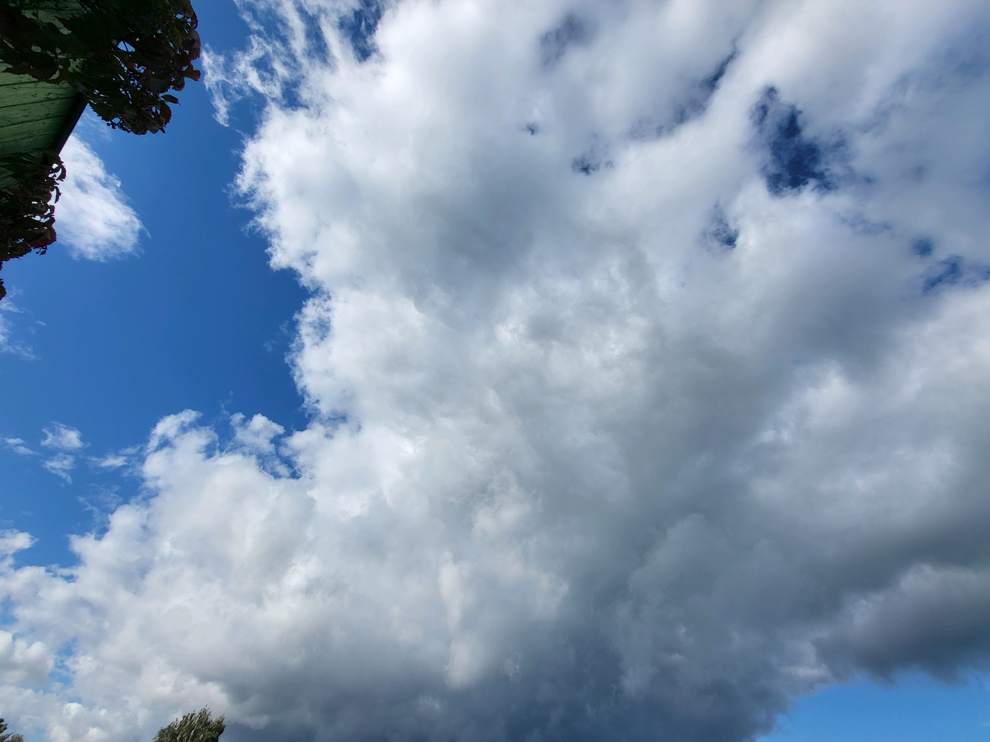 The image shows a partially cloudy sky over Ozolnieki, Latvia. In the upper left corner, a portion of a green roof and dark foliage is visible. The sky is a vibrant blue, with large, fluffy white cumulus clouds dominating the frame. Some darker, more ominous clouds are present towards the bottom of the image, suggesting a change in weather. The overall scene depicts a natural outdoor environment, likely during the daytime. There are no people or specific actions depicted in the image.