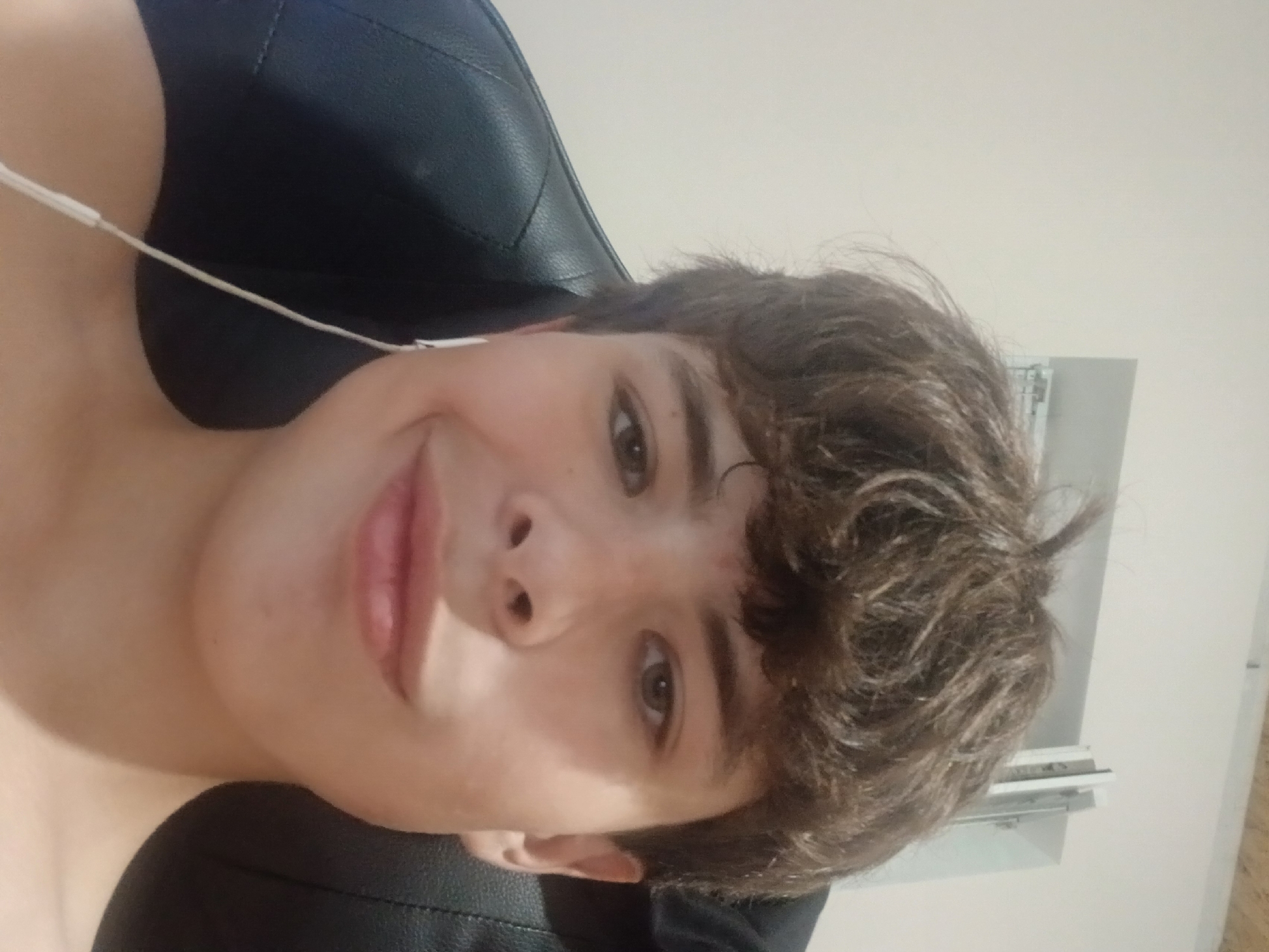 A young person with brown, messy hair and brown eyes is looking directly at the camera, wearing white earbuds. They are leaning back in a black chair, with a plain, light-colored wall behind them. A window frame with some metal objects is visible in the background. The person's shoulders and upper chest are visible, and they are not wearing a shirt. The lighting is soft and appears to be coming from the front. The image was taken indoors, likely in a residential or office setting, given the wall and window. There are no discernible landmarks or features that would definitively place this image in Dnipro, Ukraine, or any specific city.