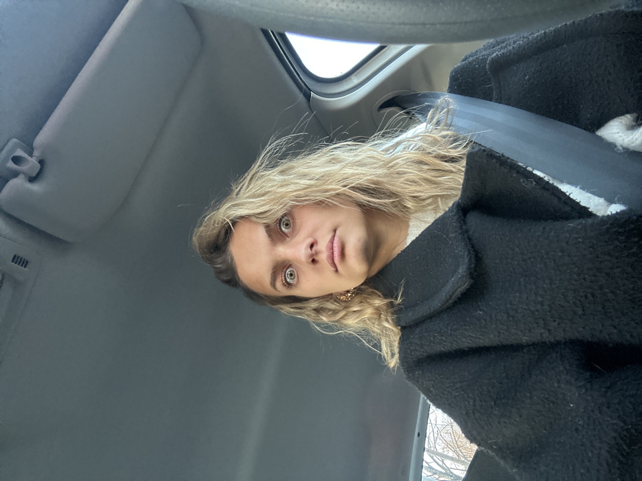 A woman with blonde, wavy hair and striking green eyes sits in a car, looking directly at the camera. She's wearing a white top and a thick, black wool coat. The car's interior is visible, including a grey sun visor and the dark grey seatbelt draped across her chest. The lighting is bright, suggesting it's daytime. The angle is slightly tilted.