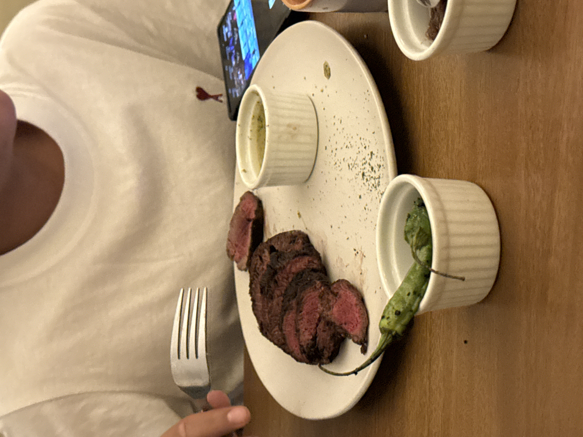 The image shows a partially eaten meal of sliced steak on a white plate, accompanied by two small ramekins, one containing a greenish sauce or condiment, and the other containing a roasted green pepper. A fork is positioned near the plate. The person eating is wearing a white t-shirt. A phone is nearby. The table appears to be made of wood. The location is Unknown, Unknown.
