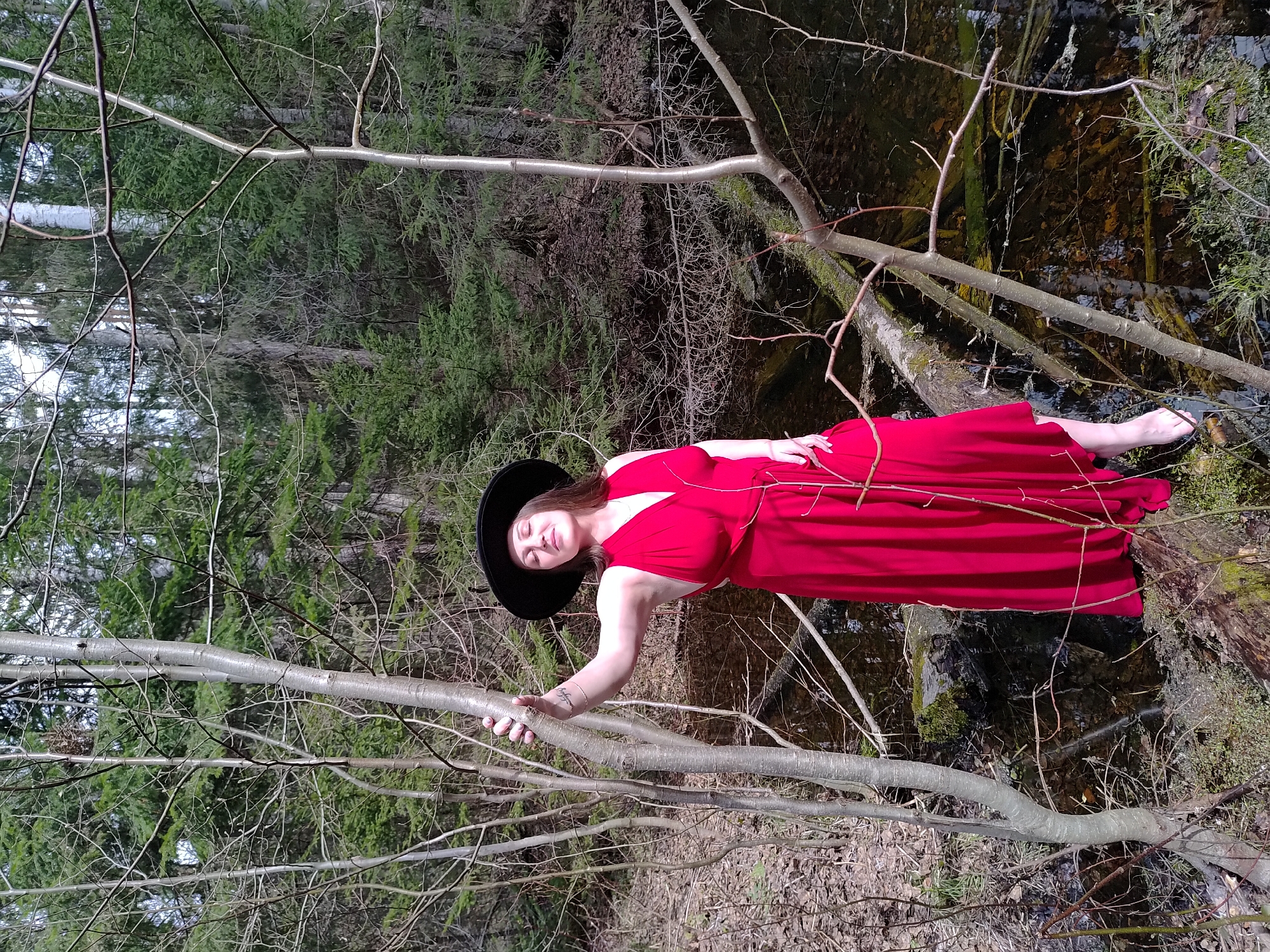 A woman in a long, red dress and a black hat is leaning on a tree branch in a forest setting. The tree branch is in the foreground and extends from the bottom left to the center of the image. The woman is positioned to the right, appearing to hold the branch with her left hand. Her dress flows down towards a body of water, possibly a stream or pond. The background features a dense forest with various trees and foliage. The location is Марковский, Russia.