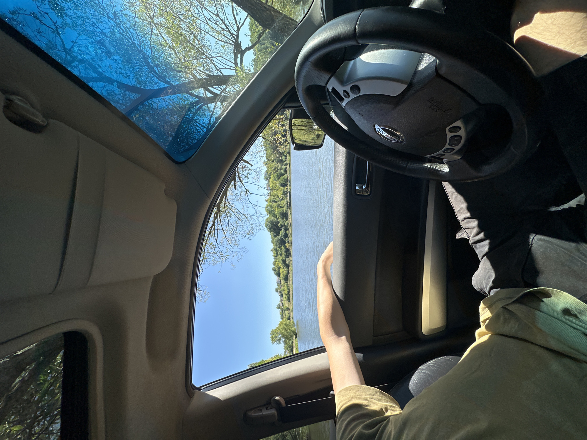 A person's arm rests on the open car door window, overlooking a body of water. The car's interior is visible, including the steering wheel and part of the dashboard. The scene appears to be a roadside view of a lake or river, with trees and a clear blue sky. The location is specified as Makiv, Ukraine.