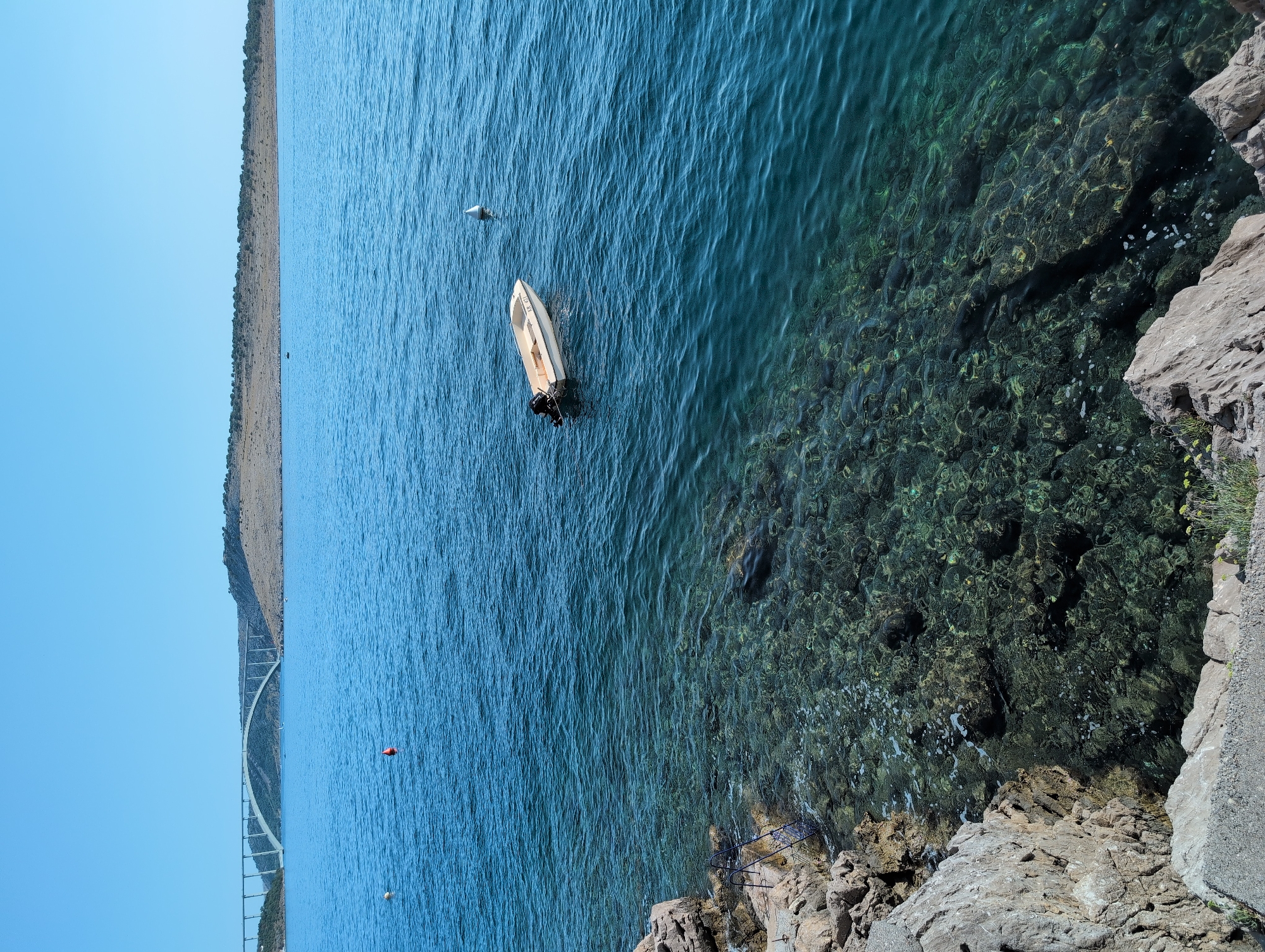 The image depicts a coastal scene in Kraljevica, Croatia, on a clear, sunny day. The foreground features rocky shoreline with a metal ladder descending into the remarkably clear, turquoise water. The seabed is visible, populated with rocks and submerged vegetation. In the mid-ground, a small, white motorboat with a black outboard motor drifts on the calm sea. Further out, a white buoy stands in the water. In the background, a long, arched bridge spans across the water, connecting landmasses. The distant shore is characterized by dry, scrub-covered hills under a bright blue sky. There are no people visible in the image.