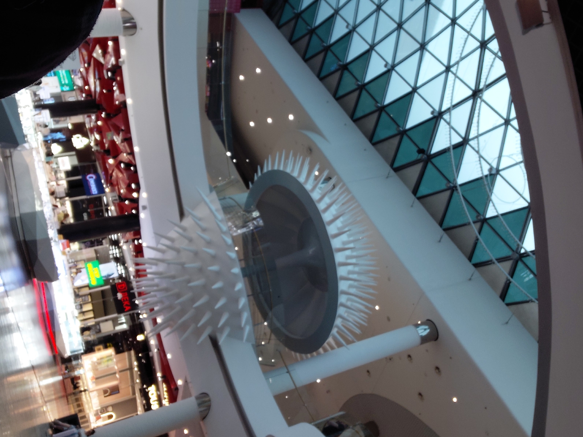 This image depicts the interior of Ocean Plaza, a large shopping mall located in Kyiv, Ukraine.
The scene captures a multi-level indoor commercial space, characterized by modern architecture. At the top, a vast, domed skylight made of numerous triangular glass panels provides natural light, with some panels appearing clear and others tinted teal. Delicate strings of small lights are suspended beneath the skylight.
Below the skylight, the upper levels feature broad, curved white walkways with glass railings and large, cylindrical white support columns, some topped with illuminated decorative elements. Recessed spotlights are visible in the smooth ceilings.
Dominating the central mid-ground is a striking, two-part artistic installation. The primary feature is a large, circular disc from which numerous white, pointed, spike-like elements protrude downwards and outwards, creating a starburst or sea urchin-like appearance. A smaller, similar spiky structure is positioned beneath it.
On the ground floor, a wide-open area with a highly reflective, polished floor is visible. Various retail stores line the perimeter, identified by signs such as "DEKA" (with a red logo), "TOUS" (with a yellow logo), and a store featuring a "ROLEX" sign. Open display cases, brightly illuminated, are arranged in the center of the floor, likely showcasing jewelry or watches. A distinct red light strip runs across the floor in the foreground. Several indistinct individuals are present, some walking, and one person is seated on the right. The dark, blurred shape in the lower-left foreground is consistent with a human shoulder or head, likely belonging to the person capturing the image.