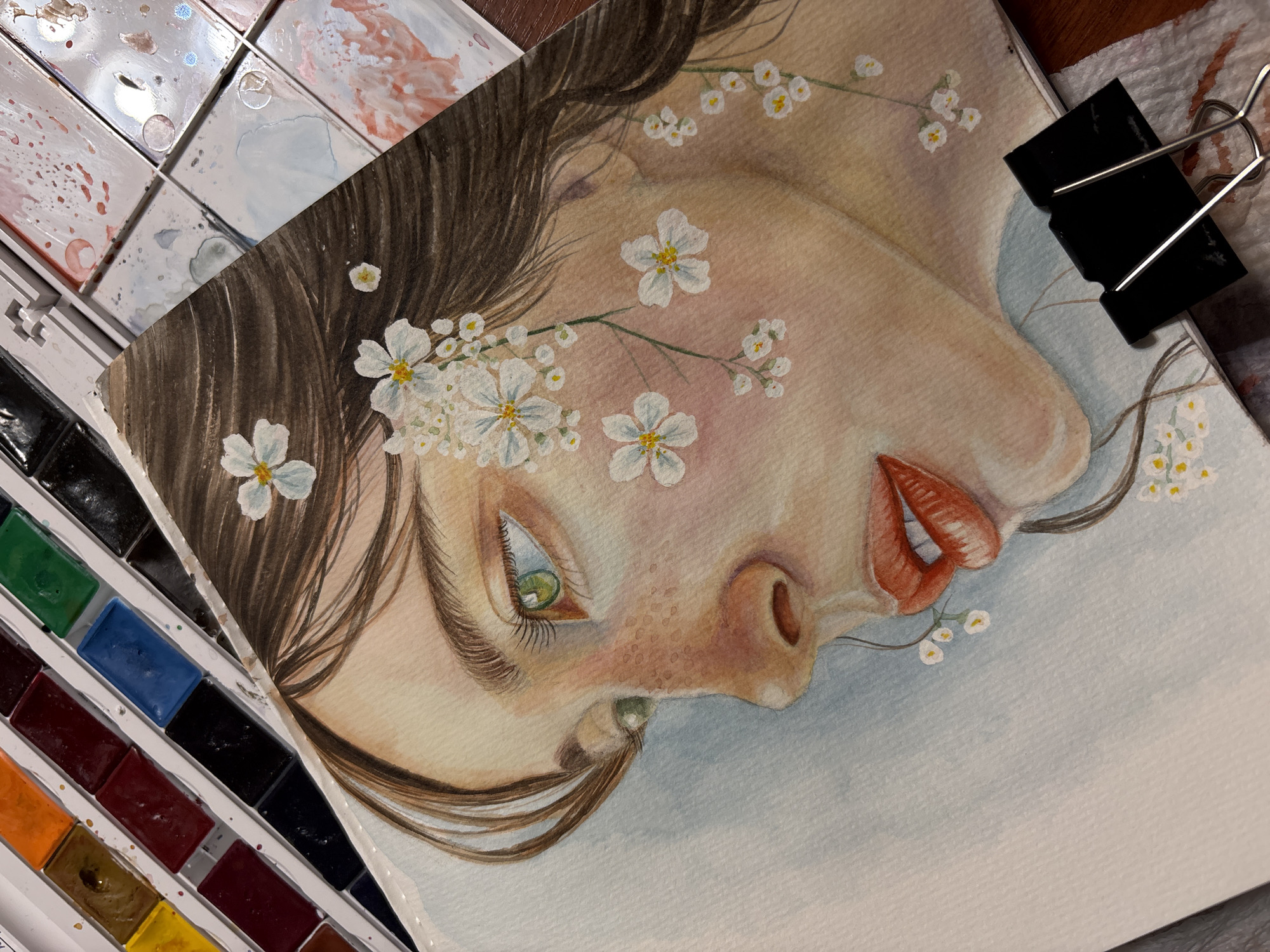 A watercolor painting of a woman's face is partially visible. The face is in profile, facing right. She has fair skin, light green eyes, and brown hair with white flowers intertwined. The lips are painted with red and orange tones. The painting is clipped to a white surface with a black binder clip. A watercolor palette is visible to the left, containing various colors. The background of the painting is a light blue wash. The location in the city of Izhevsk, Russia is unknown, as the image does not contain any information that supports this.