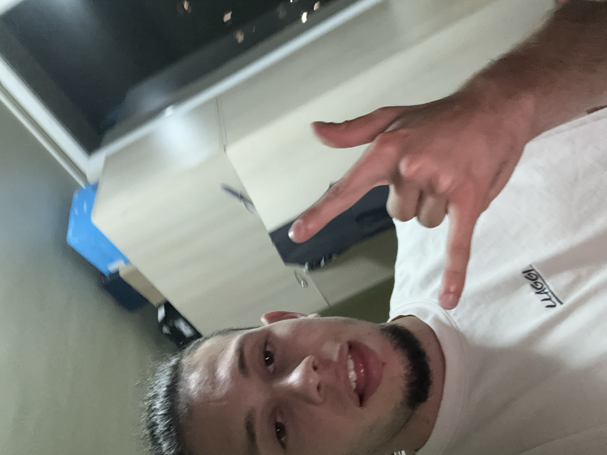 A young adult male is captured in a close-up selfie, oriented slightly to the left, with his gaze directed at the camera. He possesses dark hair pulled back from his forehead and a dark chin-strap beard. His mouth is slightly ajar, revealing his front teeth and the tip of his tongue. He wears a plain white crew-neck t-shirt featuring the dark blue, underlined text "LLAGGI" across the chest. His right hand is raised to chest height, making a gesture with the thumb and little finger extended, while the middle and ring fingers are curled.
The background is an indoor setting. To the upper left, a light green or gray wall/ceiling is visible. Behind and to the right of the individual, a light-colored, possibly beige or off-white, wardrobe or cabinet occupies the space. On top of this furniture, a bright blue rectangular object, a dark rectangular object (possibly electronic equipment), and another light-colored item are stacked. Further to the right, a large dark window is present, through which blurred, distant city lights are visible, indicating an exterior nighttime scene. There are no discernible visual cues within the image to identify the specific geographical location as the city of Stara Zagora, Bulgaria.