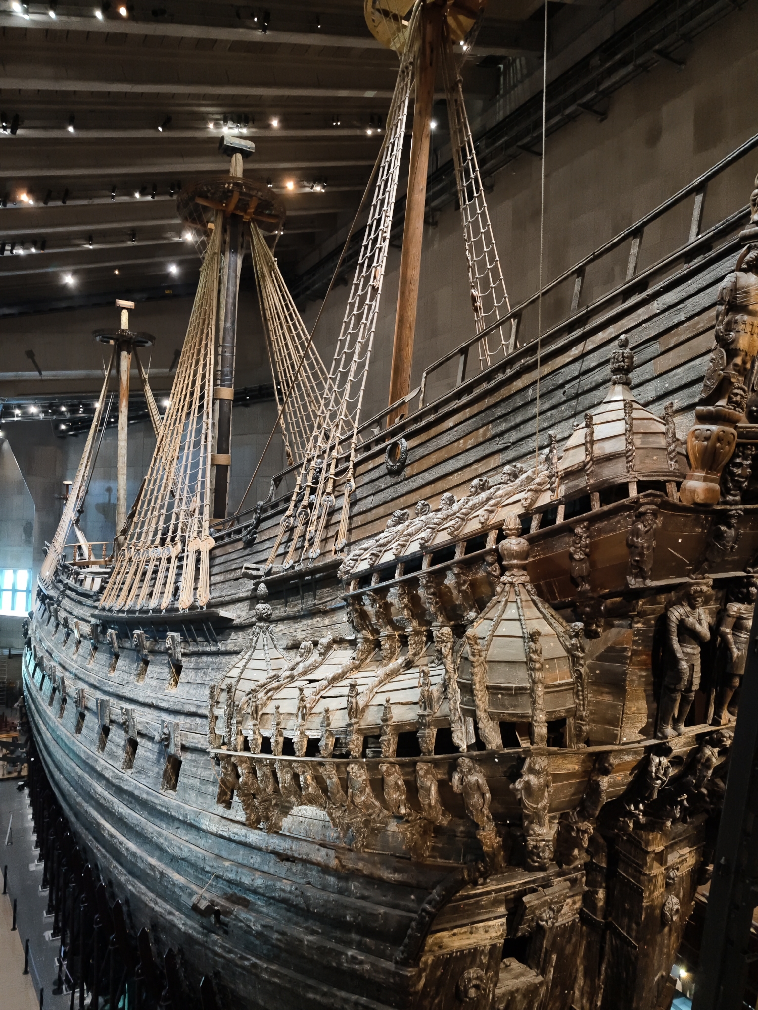 The image depicts the Vasa, a 17th-century Swedish warship, displayed indoors at the Vasa Museum in Stockholm, Sweden. The ship is heavily ornamented with intricate wooden carvings, visible along its hull and superstructure. Masts and rigging are prominent features of the ship's design. The museum environment is evident from the ceiling with visible lighting fixtures and structural elements, as well as the surrounding walls. The perspective is from a slightly elevated angle, looking towards the stern of the ship. There are no people visible in the image.