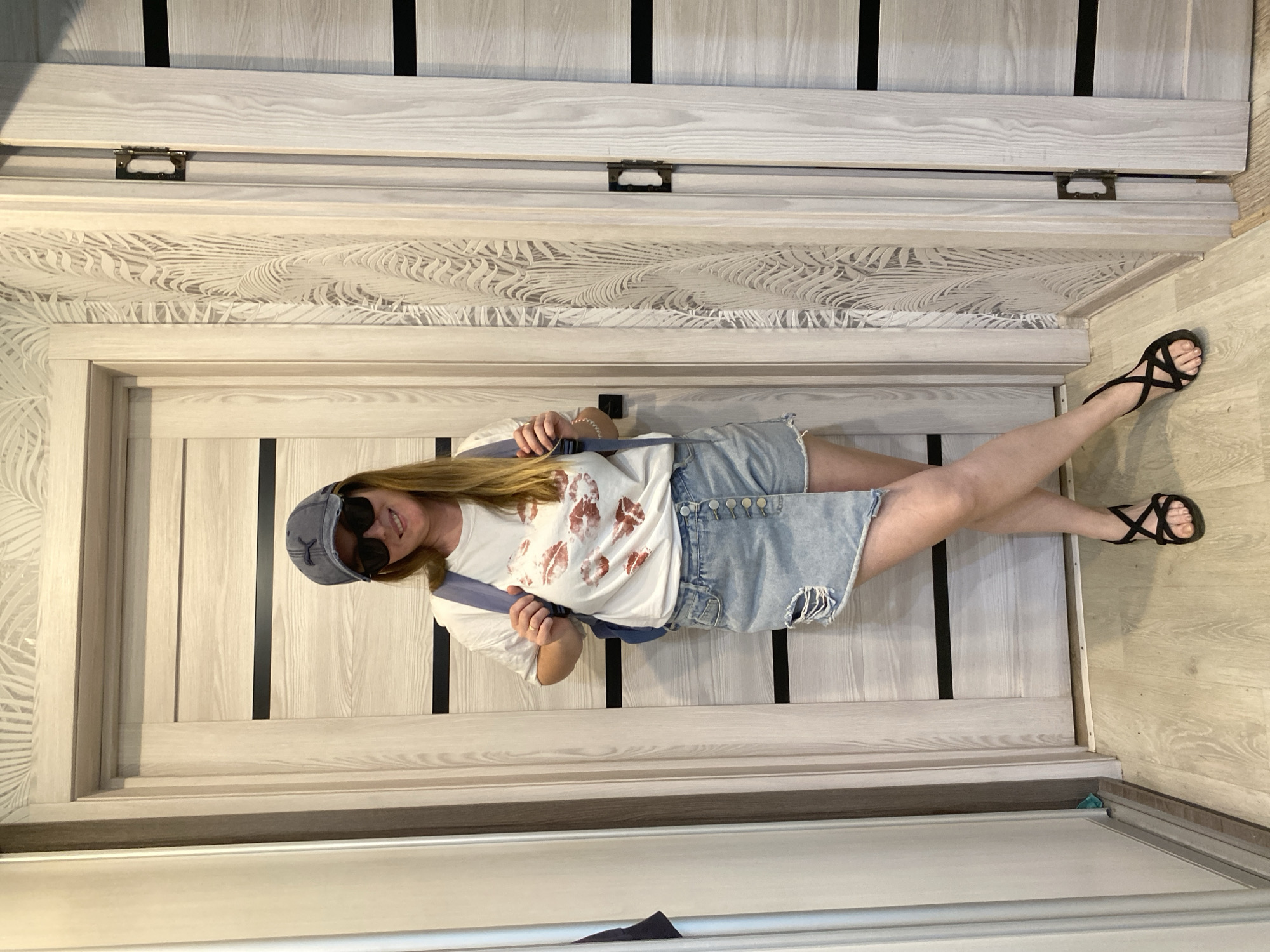 A full-body shot captures a woman inside an apartment in Noviy, Russia. She is wearing a white t-shirt with red lip prints, a denim mini skirt with a button-up side, a blue baseball cap, and sunglasses. She is also wearing black strappy sandals. The woman is positioned in front of a closed, light-colored door with dark vertical accents. A light-colored wardrobe with a decorative design is behind the door. The apartment has light-colored wood flooring. The image appears to be taken in a residential setting.