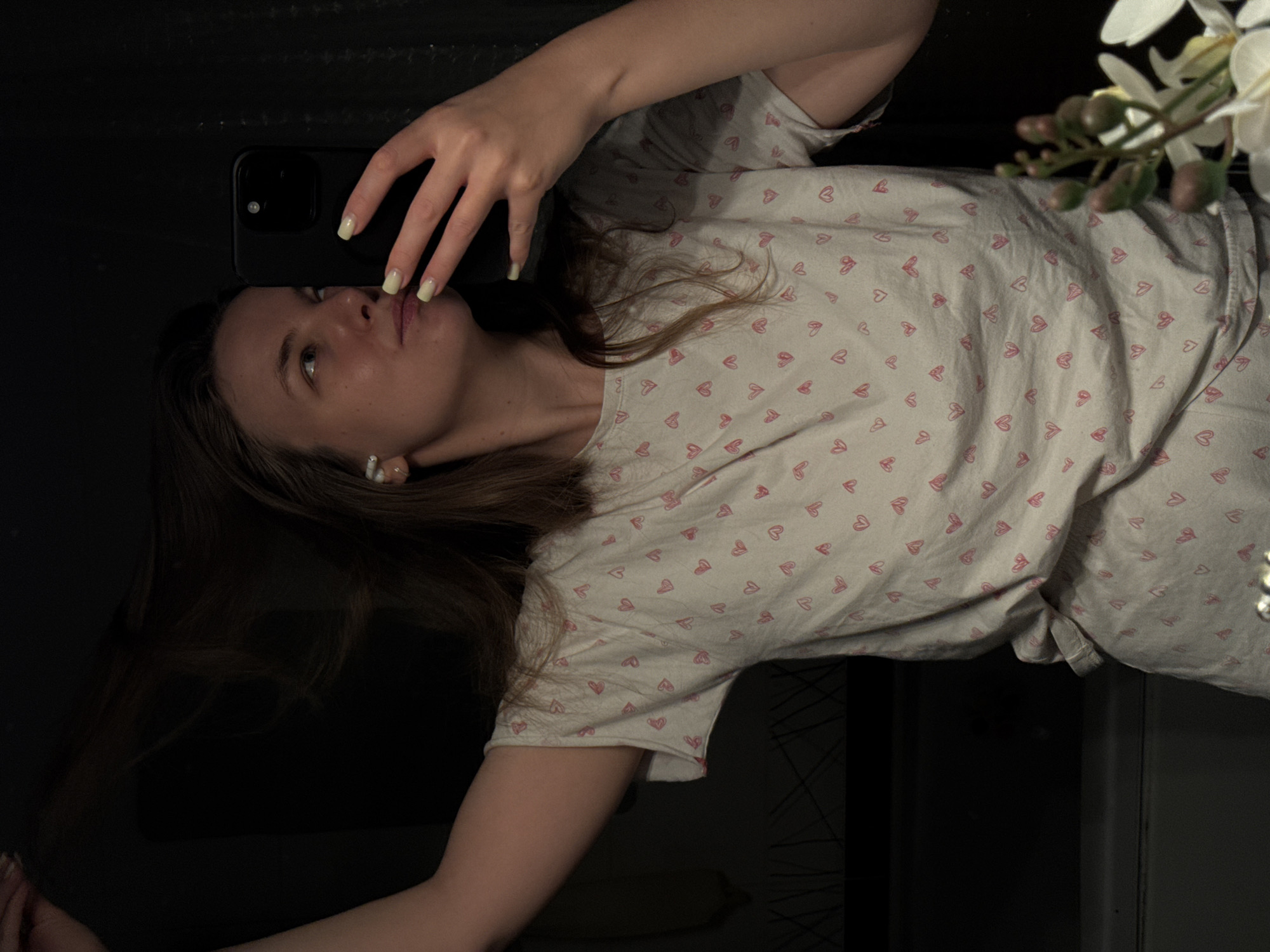 A woman lies on a dark surface, angled diagonally in the frame, taking a selfie with a black smartphone. She wears a light-colored t-shirt patterned with small red hearts. Her head is tilted back, her face visible, and her hand holds the phone up. A flower arrangement is in the upper right corner. The location in the city of Vladimir, Russia, cannot be determined from this image.