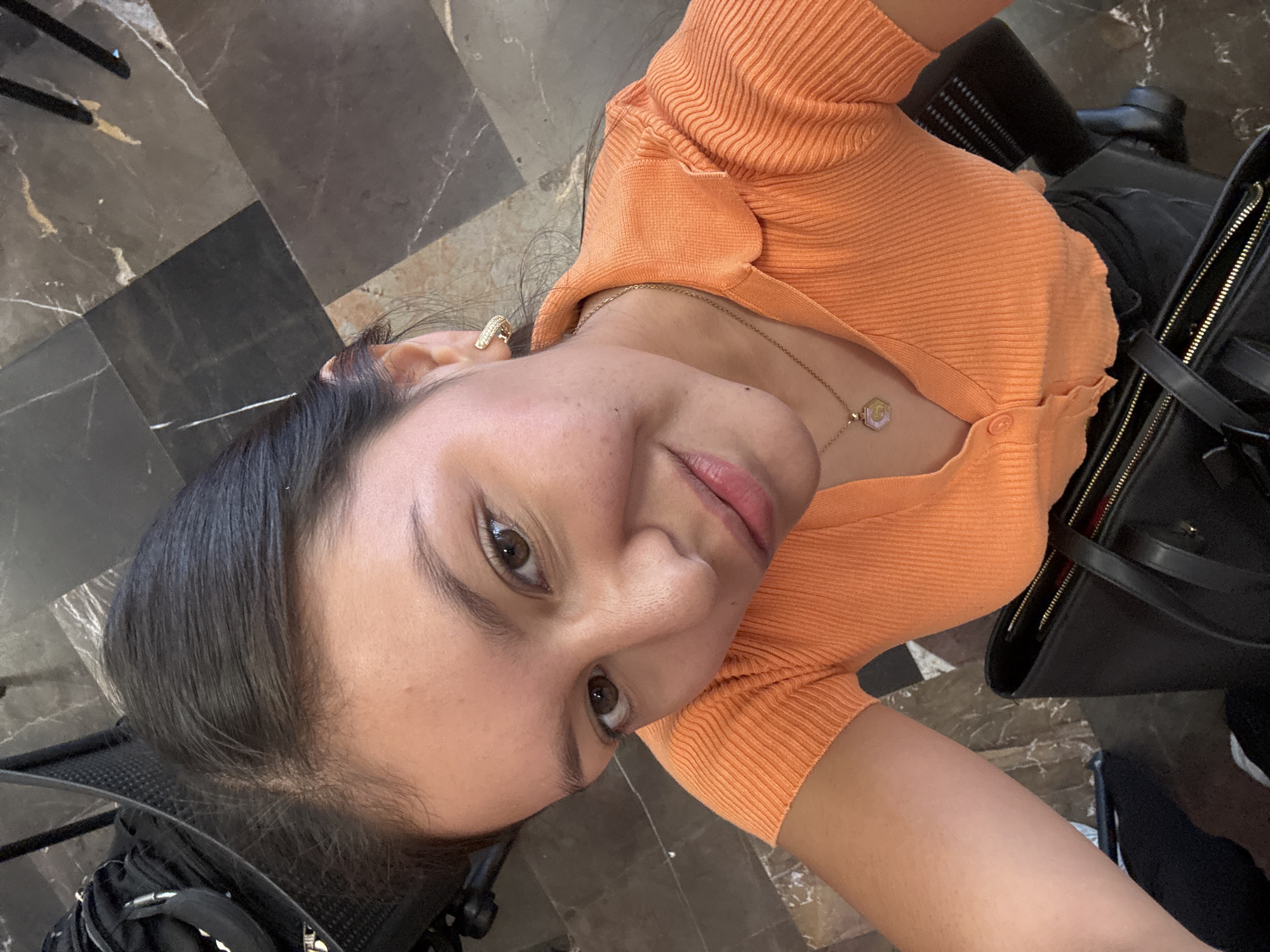 A medium-shot selfie photograph depicts a young woman with olive skin. She has dark hair and brown eyes. She is wearing a ribbed, orange, button-up short-sleeved top and a gold necklace with a hexagonal pendant. Her left ear has a square-shaped gold earring. To the left is a black bag and partially visible black chair. The background features a dark, marble-tiled floor with light veining. The location is most likely in San Andrés Cholula, Mexico.