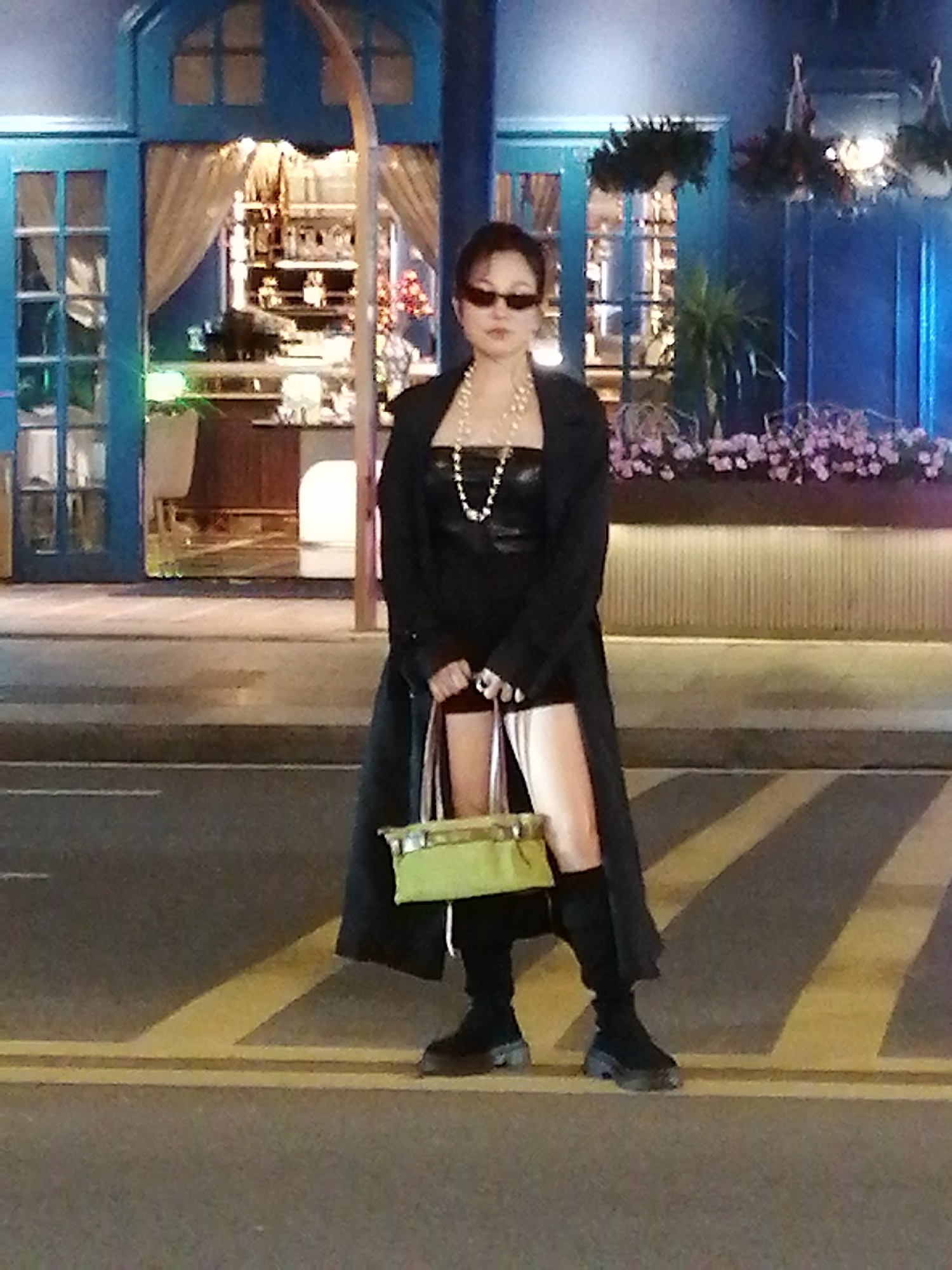 A woman stands on a crosswalk in front of a building with a blue facade in Da Lat, Vietnam. She is wearing a black coat over a black top, a pearl necklace, sunglasses, and black boots. She is holding a light green handbag. The building has large windows and ornate trim, and appears to be a restaurant or bar, with plants and decorative lighting visible inside and outside. The street is paved with asphalt and marked with yellow and white lines. The lighting suggests it is nighttime or dusk.