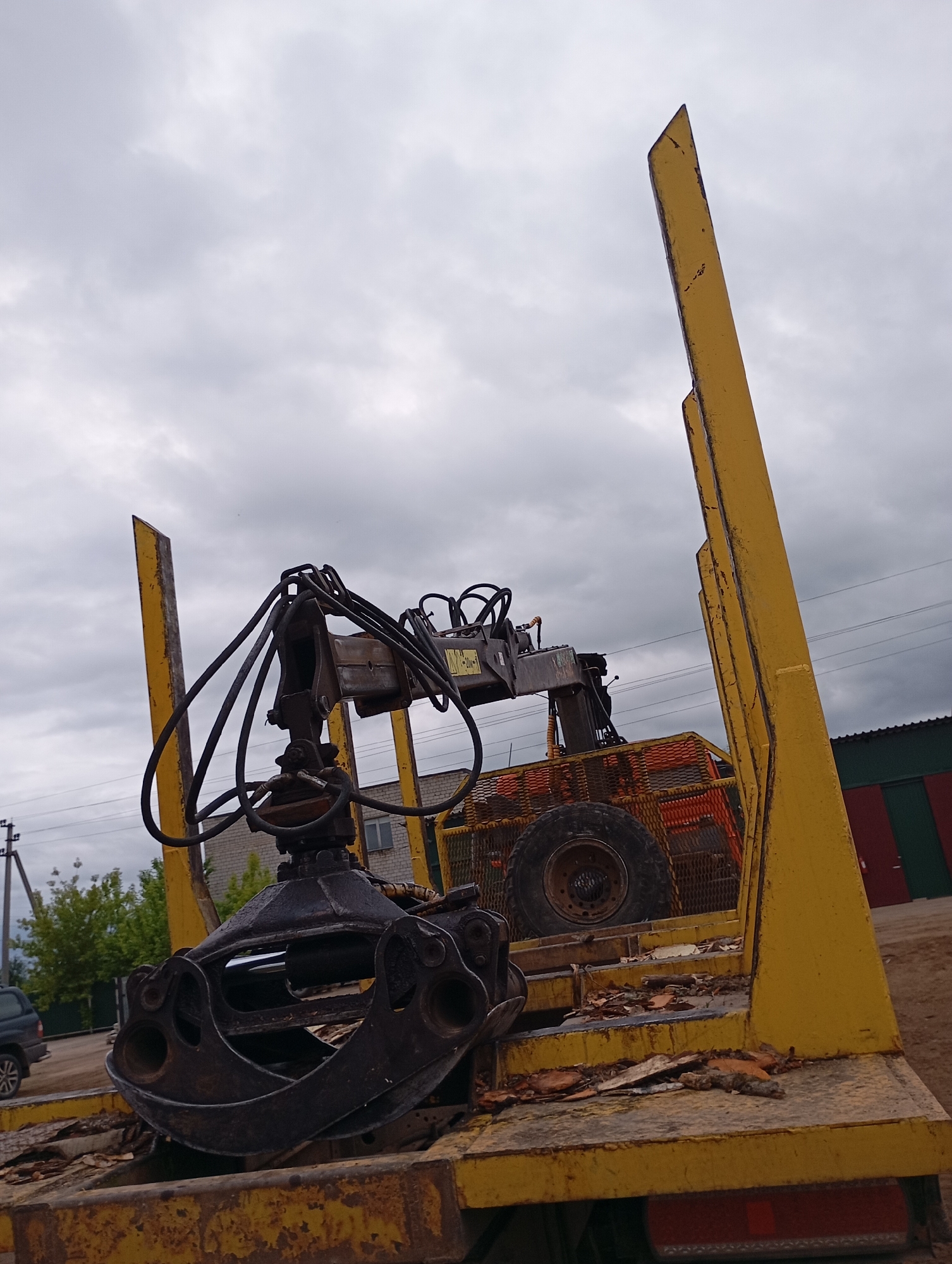 The image captures an industrial area in the city of Фоки, Russia, under a heavily overcast sky. The foreground is dominated by a weathered, yellow metal log trailer or truck bed featuring tall, pointed vertical stanchions on its sides, designed for log retention. The surface of the bed is dirty, exhibiting rust and scattered wood debris and dry leaves. A dark, heavy-duty hydraulic crane assembly is mounted on the front section of the bed, extending a large, black metal grapple over the trailer. Visible hydraulic hoses and cylinders are attached to the crane and grapple. In the background, to the left, a portion of a dark-colored vehicle is parked near a utility pole and dense green foliage. A light-colored brick building with a visible window stands in the mid-ground, with power lines running horizontally across the upper frame. To the far right, a section of a building with vertical red and dark green panels is partially visible. The ground appears to be unpaved dirt. No individuals are present in the scene.