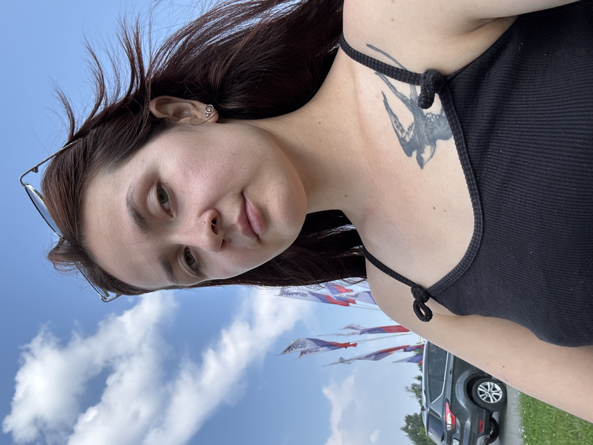 The image shows a young woman taking a selfie outdoors. She has long, dark brown hair, a nose ring, and a tattoo of a bird on her chest. She is wearing a black tank top. In the background, there are several flags flying, and a dark-colored SUV is parked on a grassy area. The sky is blue with scattered clouds. The flags appear to be Russian flags, suggesting the location may be in Russia. Without further information, it is impossible to pinpoint the exact location within Naberezhnye Chelny, Russia. However, the presence of flags and a car suggests an outdoor public space, possibly a park, event venue, or a roadside area. The overall scene is casual and appears to be taken on a sunny day.