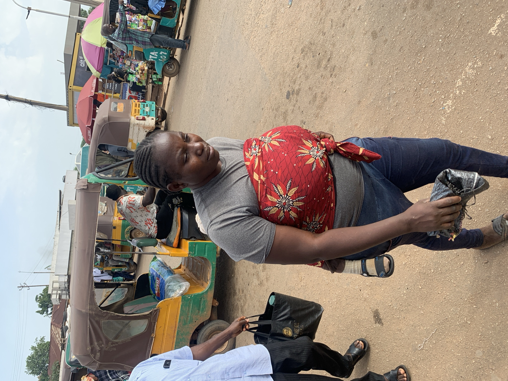 In Gura Top, Nigeria, a woman with braided hair, wearing a grey t-shirt and blue jeans, carries a child wrapped in a red and white patterned cloth around her chest. She holds a black bag in her right hand. To her left, a person in a blue and white striped shirt walks with a black shopping bag. In the background, several auto-rickshaws, also known as "Keke Napep" in Nigeria, are parked. Some of the Keke Napep vehicles are brightly colored with yellow and green, and one has a brown canopy. The scene appears to be a busy street or market area, with the ground covered in a reddish-brown dirt. Overhead, utility poles and wires are visible against a cloudy sky. Several vendors are present, with stalls displaying various goods, including umbrellas and packaged items.
