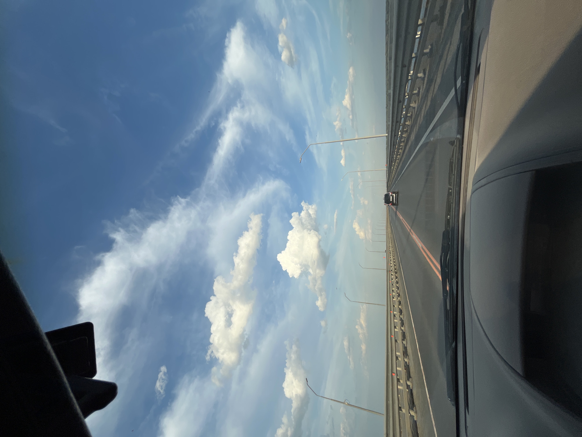 The image shows a view from the interior of a vehicle driving on a long, multi-lane bridge. The bridge has a metal guardrail along the right side, with streetlights positioned at intervals. There are lane markings on the asphalt road, including a double yellow line in the center and a white line to the right of the lanes. In the distance, a dark-colored SUV is traveling in the same direction. The sky above is a vibrant blue with scattered white clouds, some of which are wispy and others are more cumulus-shaped. The sun appears to be casting a warm light, creating shadows on the dashboard of the vehicle in the foreground. The location is identified as a bridge in or near Pristannoye, Russia.