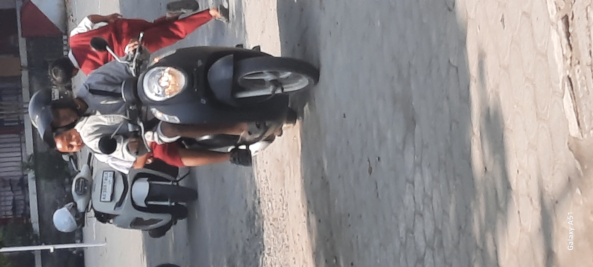 This image depicts two children riding a black scooter in Tangkil, Indonesia. The child in front is wearing a helmet and a grey jacket, with red shorts. The child behind them is wearing a white shirt and red shorts. A white scooter is parked to the left of the black scooter. The ground appears to be paved with rough asphalt and some cobblestones. Shadows from nearby structures are cast on the ground. The lighting suggests it is daytime.